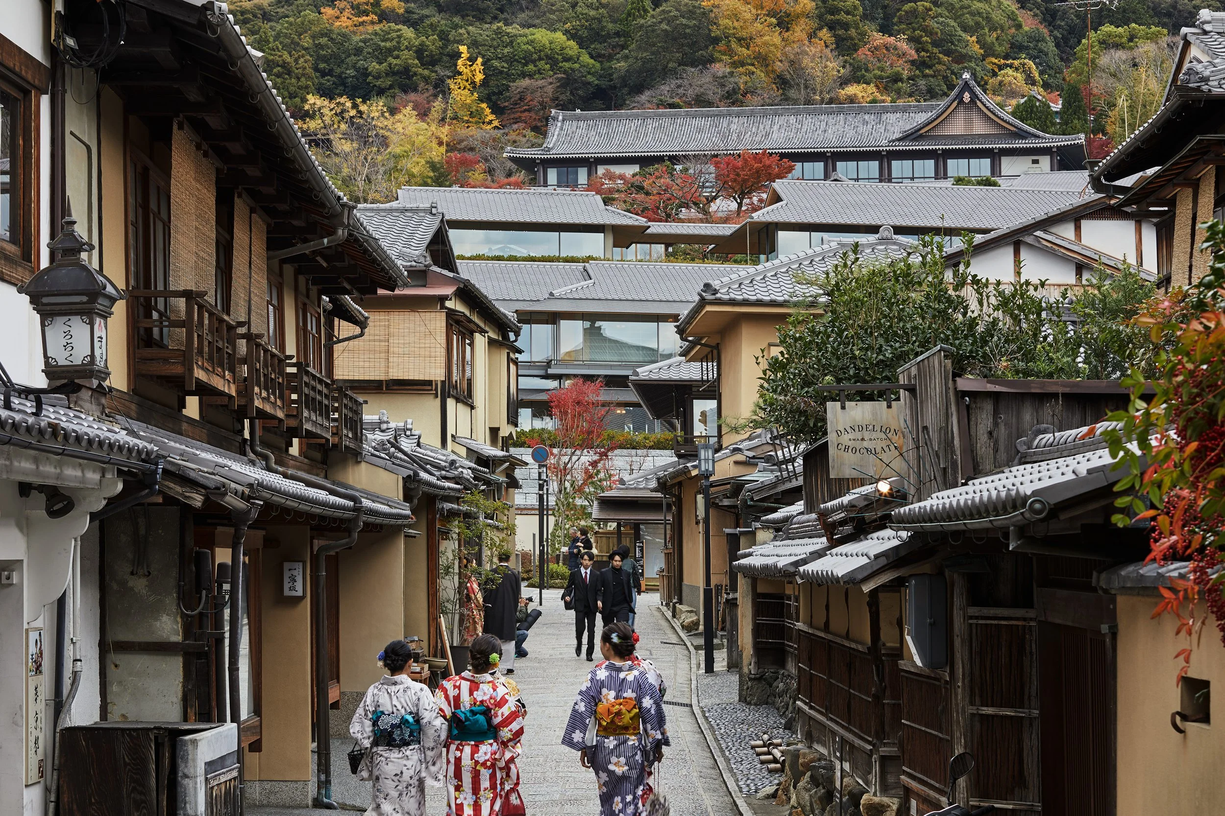 People wearing traditional Japanese kimonos walking through a narrow street with traditional Japanese buildings, trees with autumn foliage, and a wooded hillside in the background.