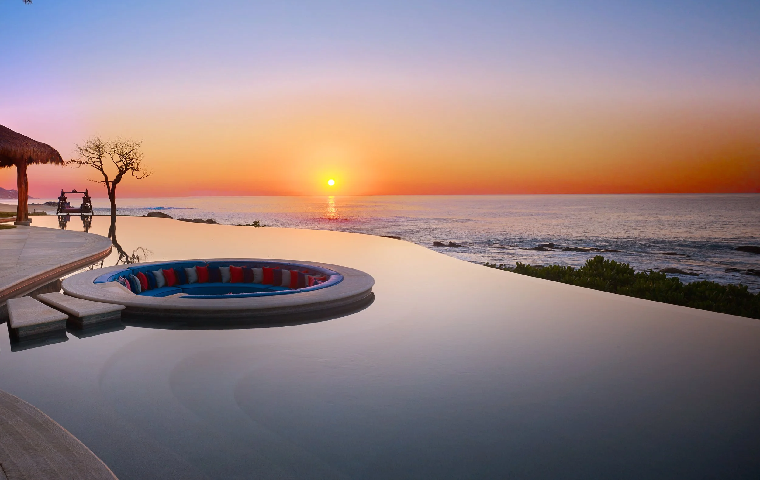 Sunset over the ocean viewed from a luxurious outdoor pool area with a circular seating nook with red and blue cushions, a thatched-roof structure, and rocky shoreline.