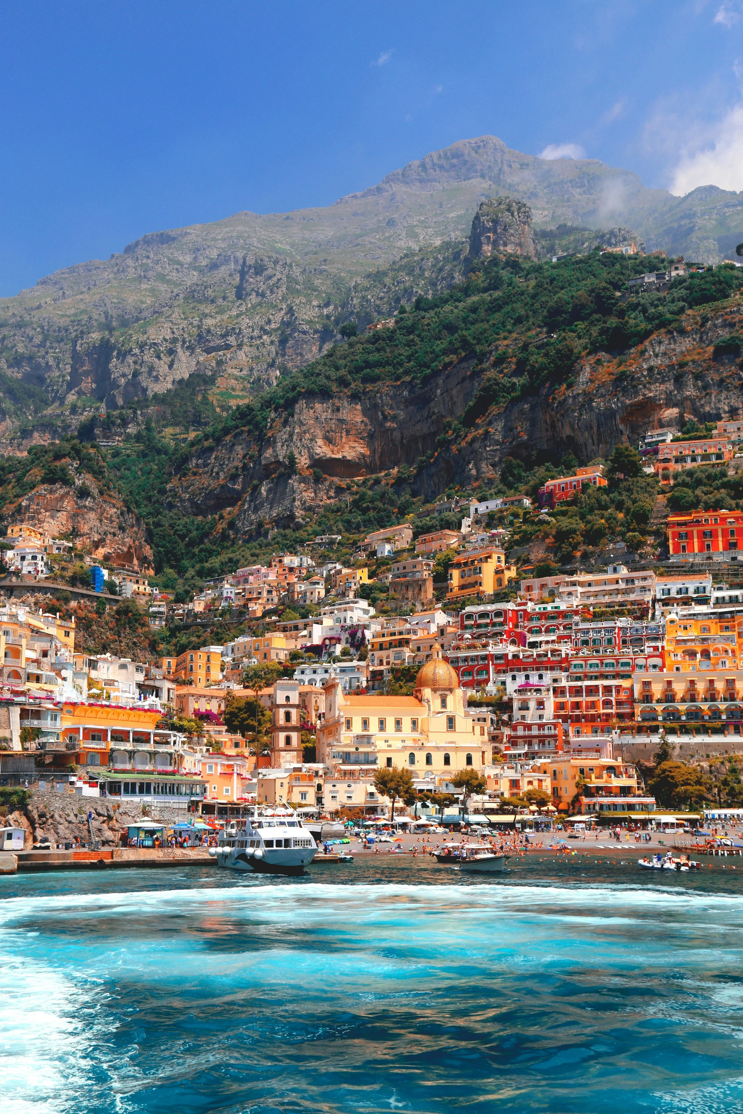 Colorful coastal town built on a hillside with boats in the harbor at the bottom and mountains in the background under a partly cloudy sky.
