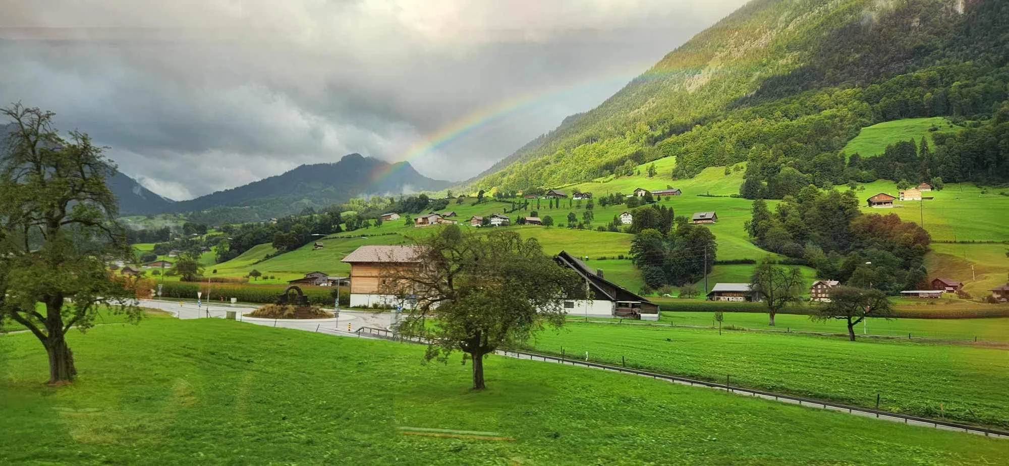A scenic green landscape in Switzerland with rolling hills, scattered trees, small houses, and a rainbow in the cloudy sky.