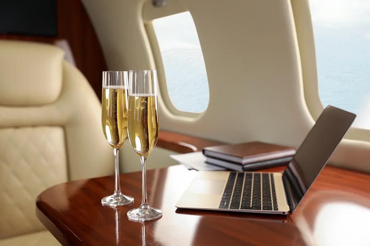 Two glasses of champagne on a wooden table inside a plane with a laptop and closed books nearby.