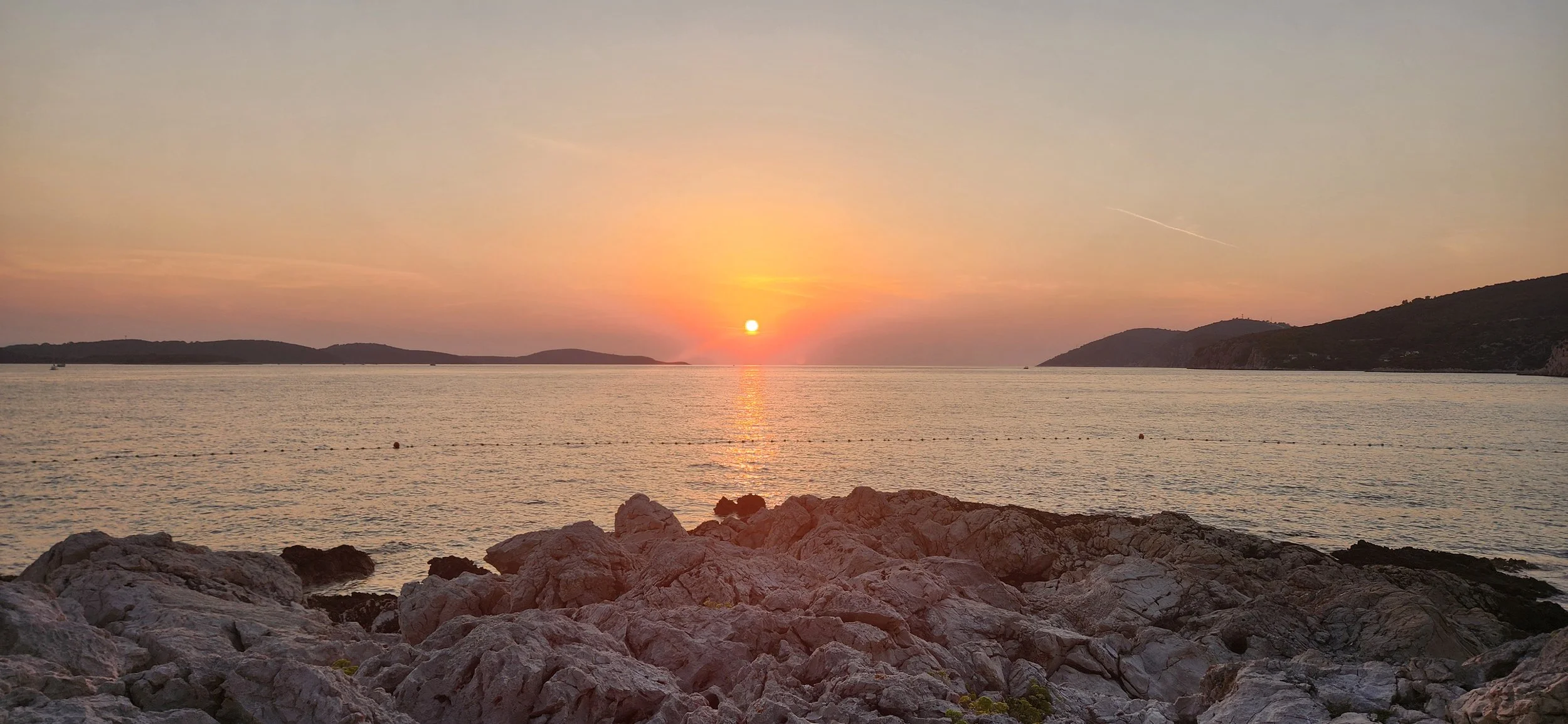 Sunset over a calm body of water in Hvar, Croatia with rocky shoreline in the foreground, cliffs and islands in the distance.