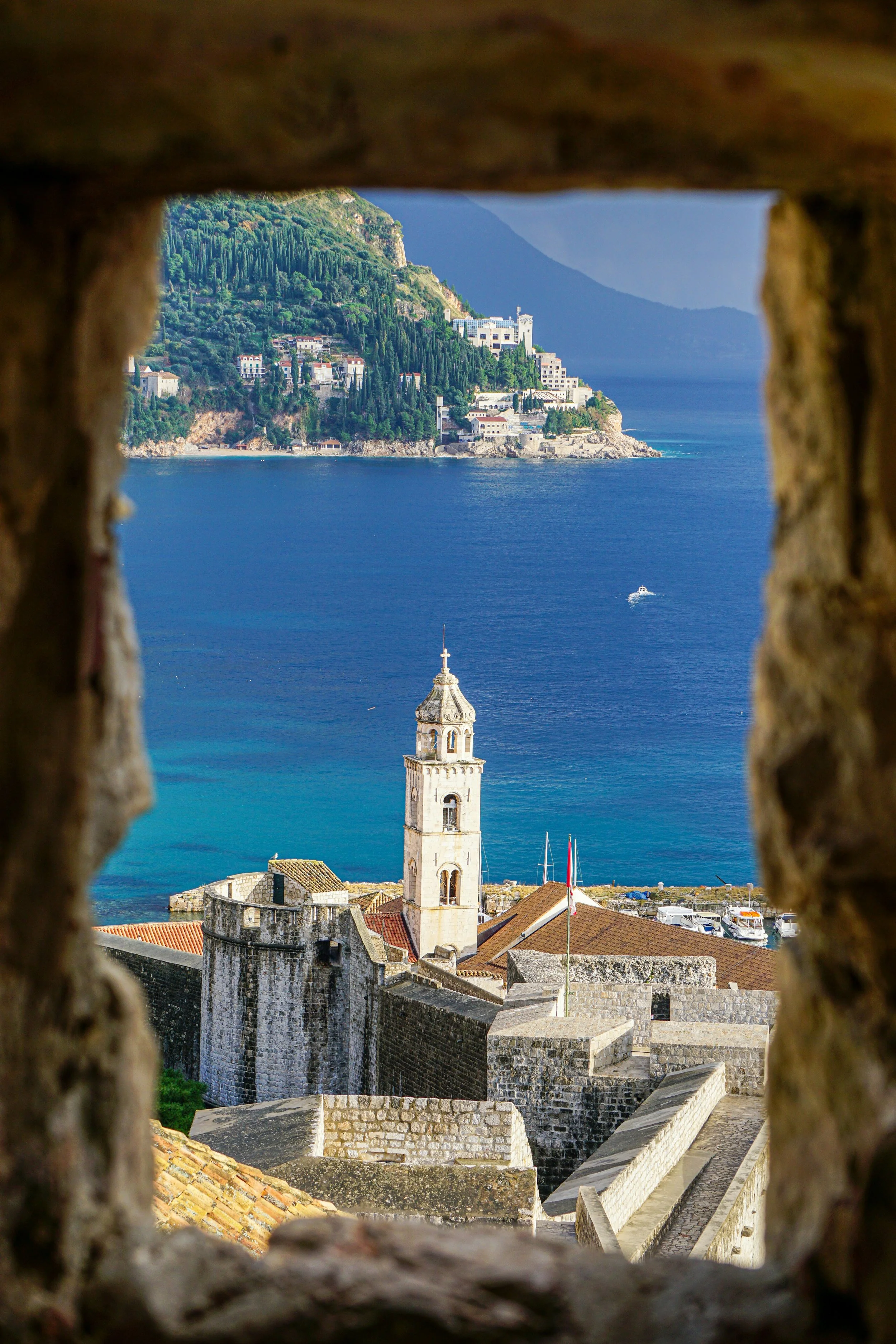 View of a historic stone fortress with a church tower, overlooking the sea and a hillside with green trees and buildings, seen through a small window in a stone wall.