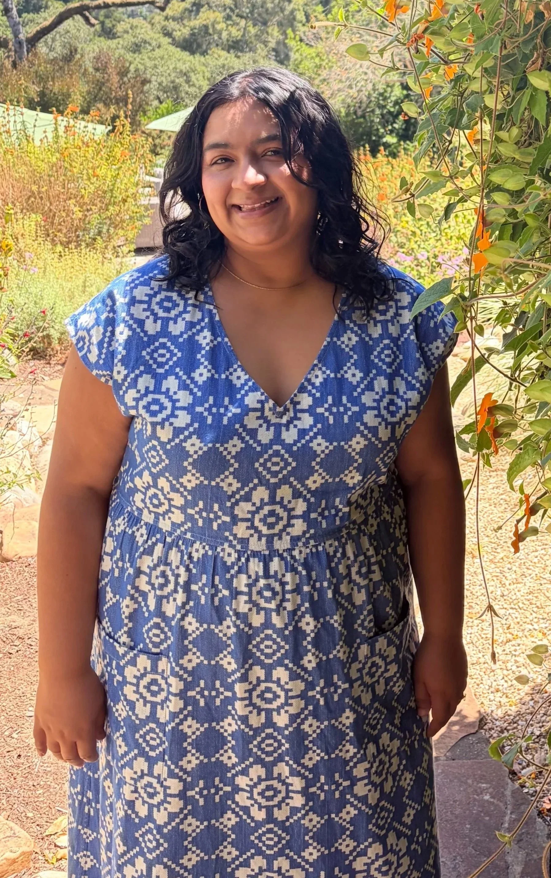 A woman wearing a blue and white patterned dress smiling outdoors with greenery and flowers in the background.