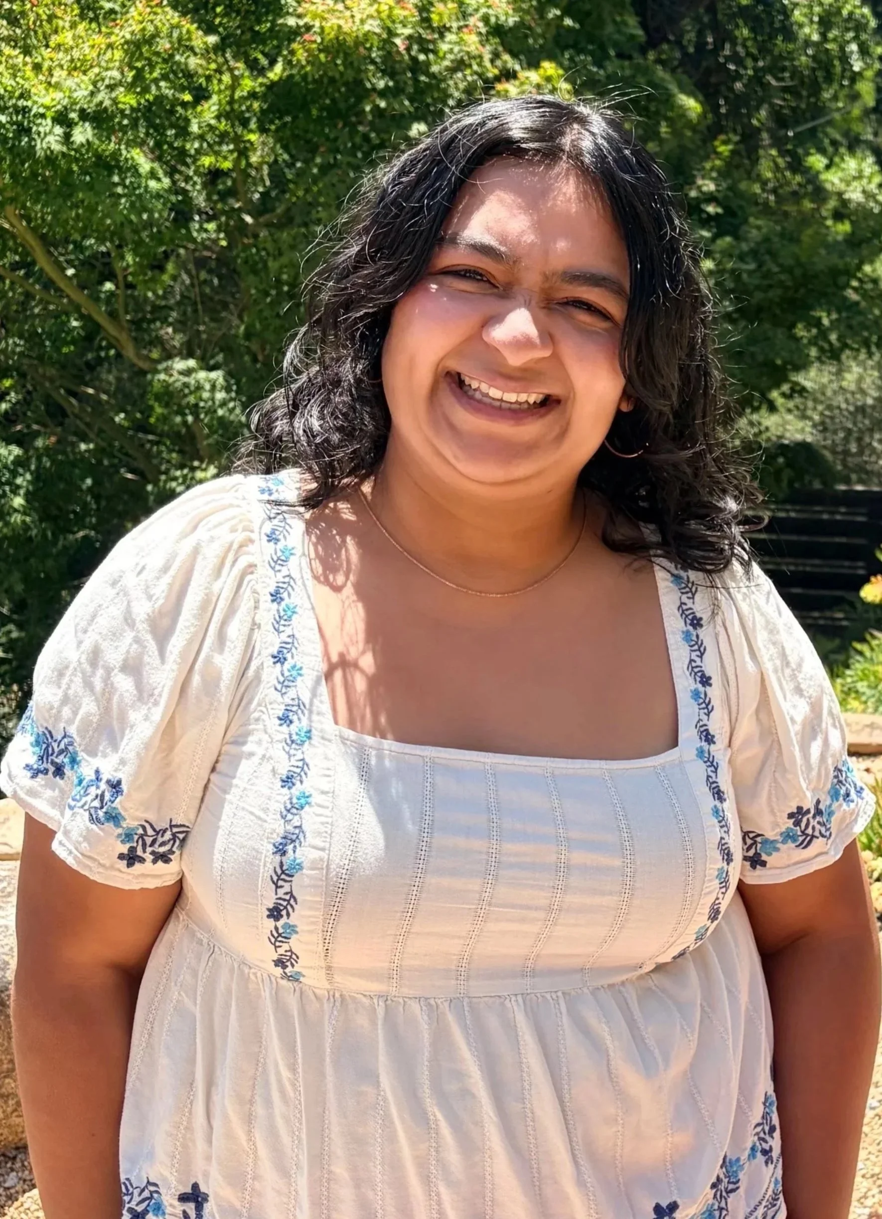 Close-up photo of a woman with curly black hair, smiling outdoors with green trees in the background, wearing a cream-colored blouse with blue embroidery.
