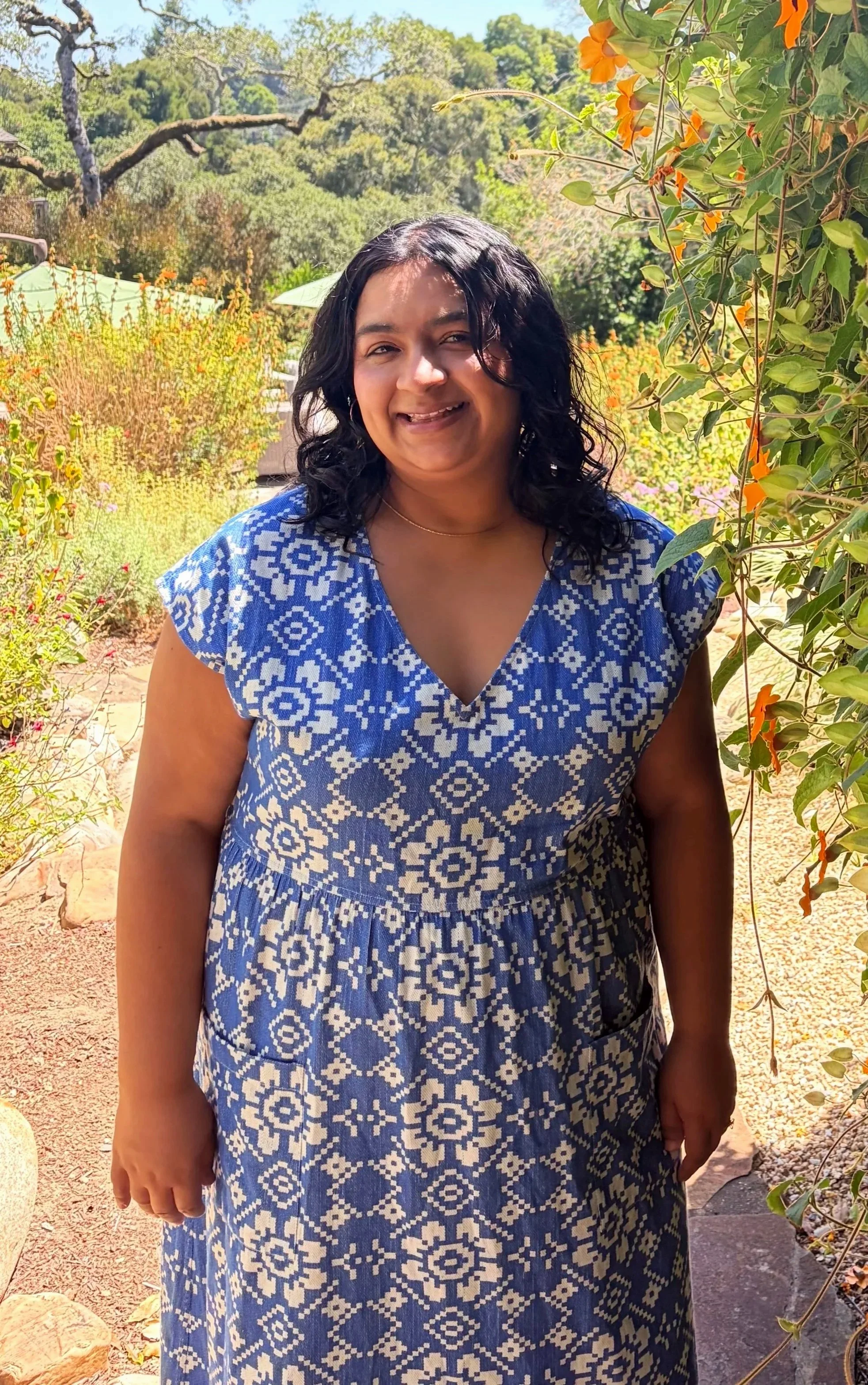 A woman with dark hair wearing a blue and white patterned dress standing outdoors in a garden with trees and plants.