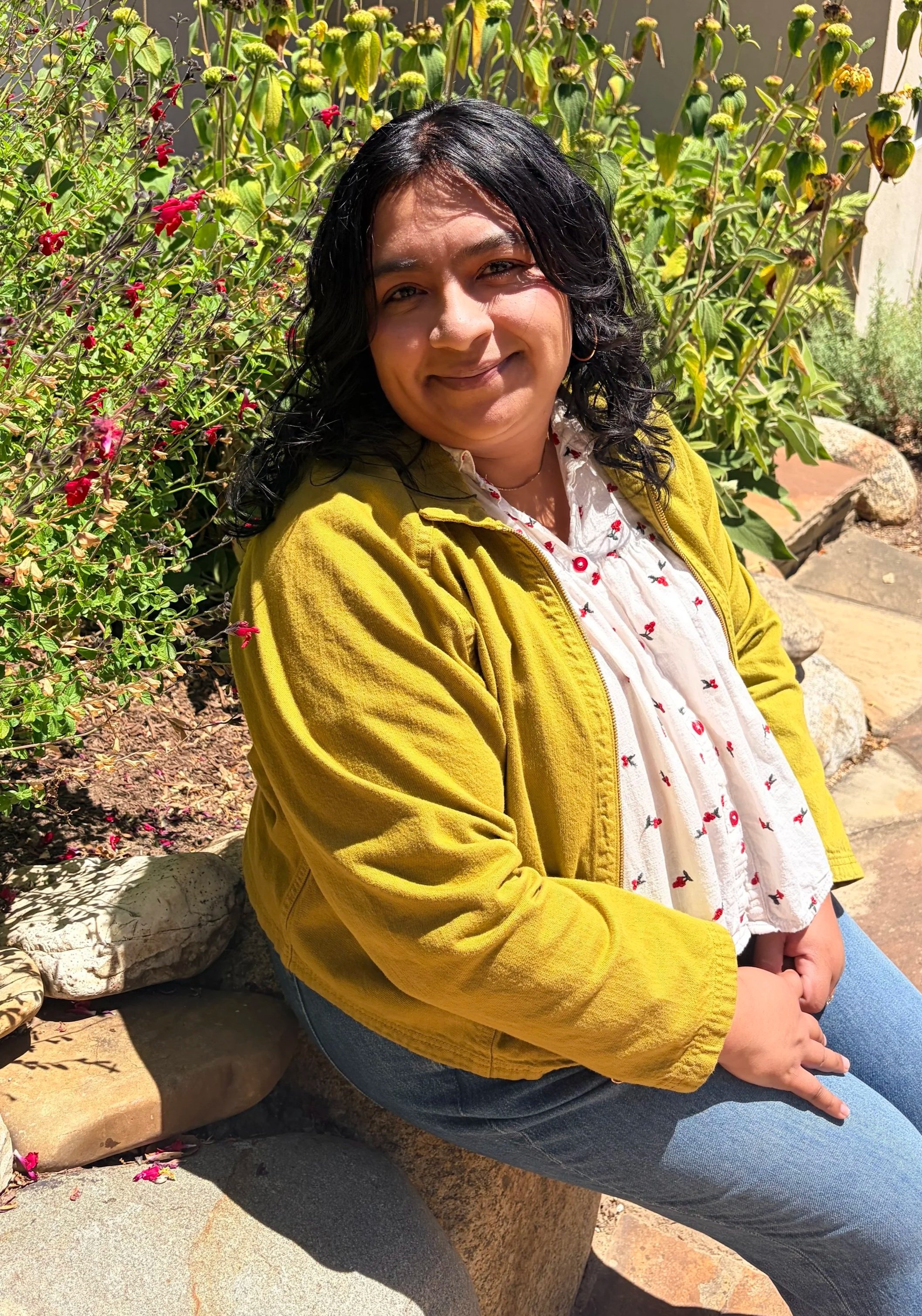 A woman with black curly hair and a smile, sitting outdoors among plants and rocks, wearing a yellow jacket over a white shirt with a red and black pattern, and blue jeans.