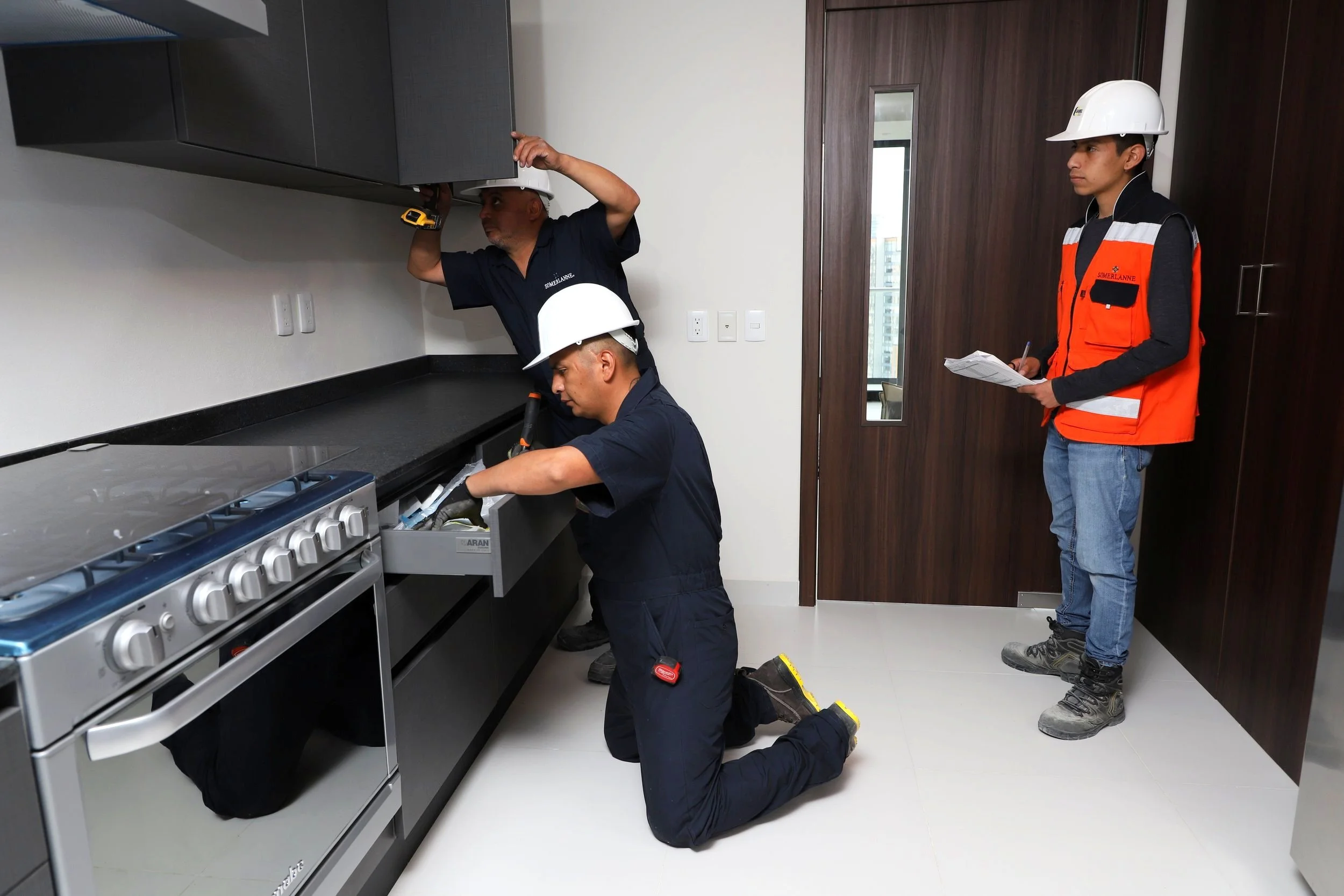 Tres trabajadores Somerlanne, inspeccionando una cocina, uno tomando notas, otro revisando un gabinete y el tercero de rodillas revisando un cajón. Usan cascos blancos de seguridad y uniformes de trabajo. Se observa una estufa y gabinetes grises.