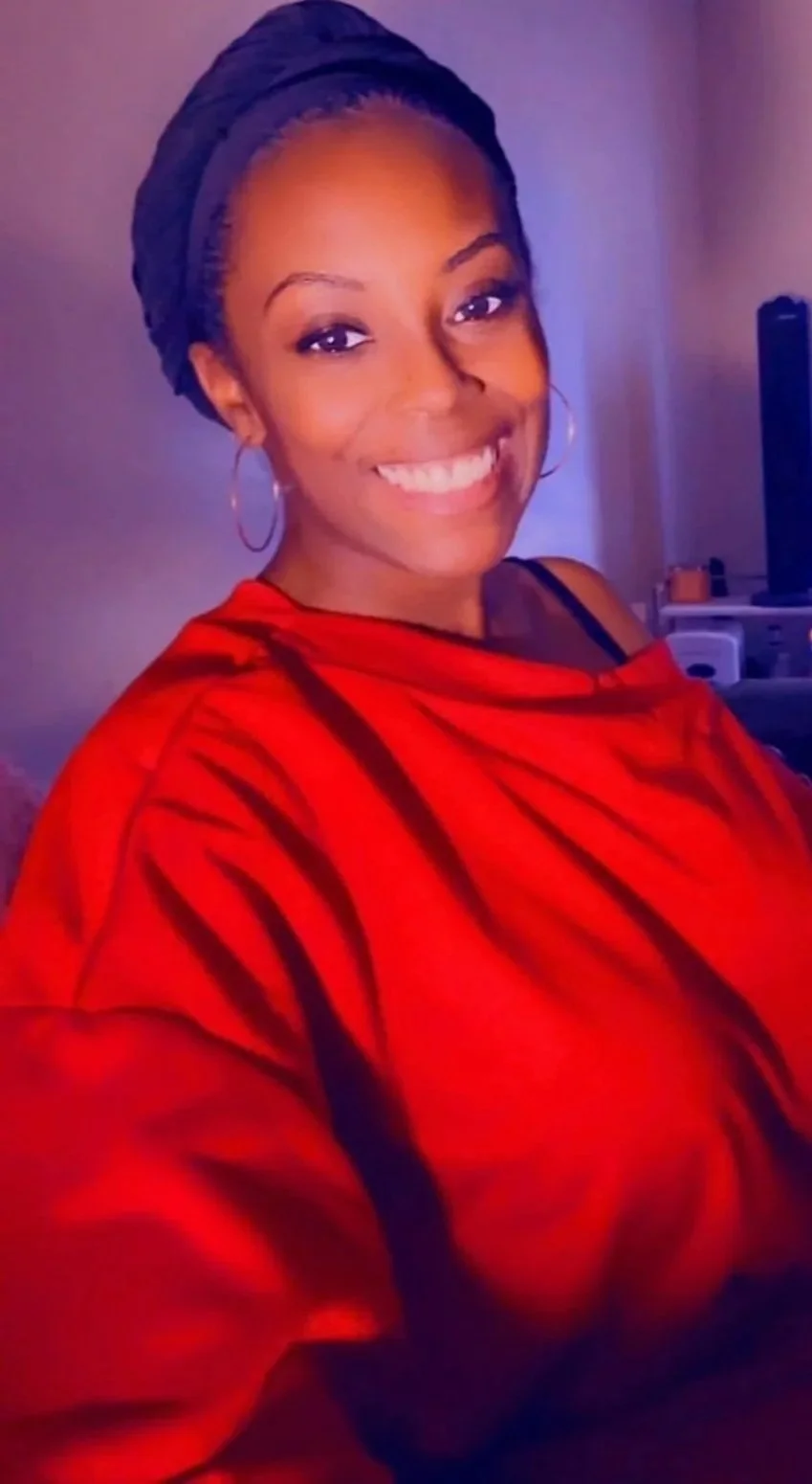 A young woman with a broad smile, wearing a black head wrap, hoop earrings, and a red top, sitting in a room with a television and some furniture in the background.