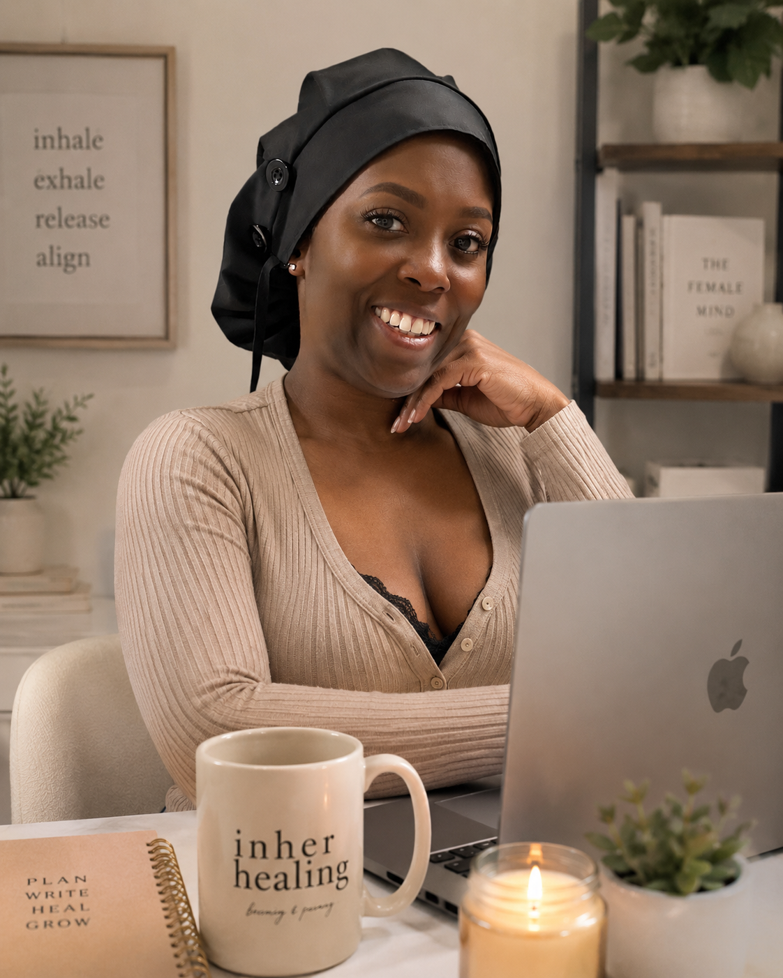 A woman wearing a black hair bonnet, smiling and sitting at a desk with a laptop, a mug that says 'inner healing,' a journal, a candle, and a small plant, in a cozy room with books and framed art on the wall.