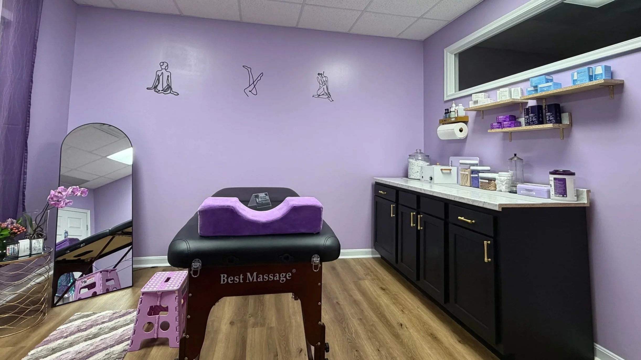 A massage therapy room with purple walls, black cabinets, wooden shelves with skincare products, and artwork of line drawings of women in yoga poses on the wall. There is a massage table with purple cushioning, a mirror, flowers, and various supplies.