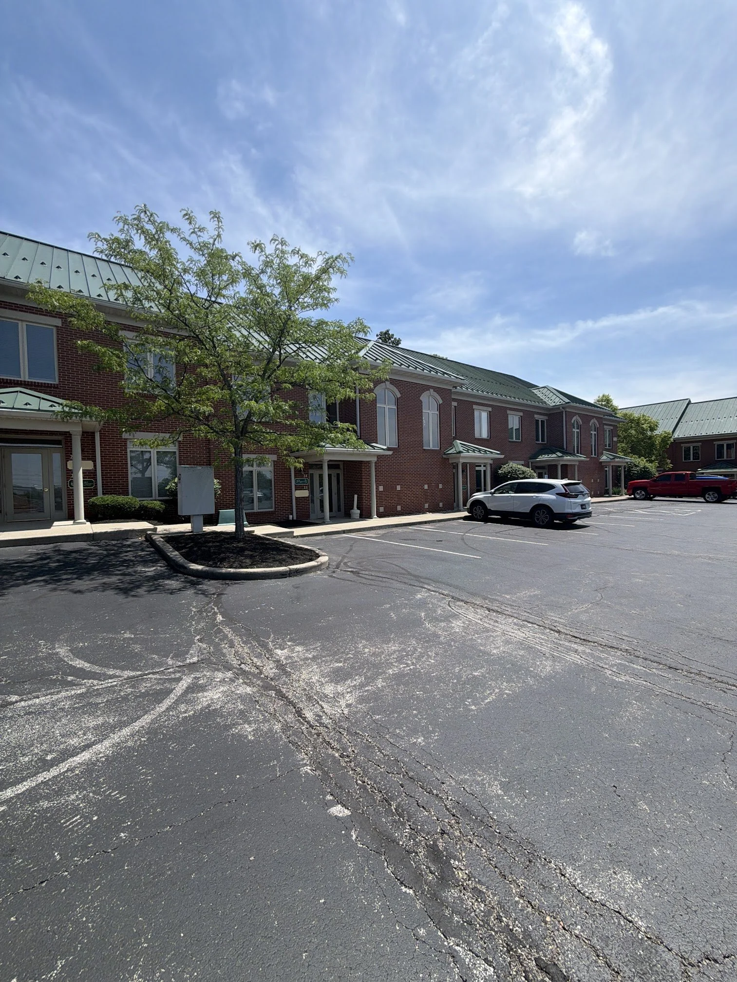 View of a parking lot with a few cars, a brick building with multiple windows, a tree, and a partly cloudy blue sky. This image gives a view of where The Purple Wax Suite is located when looking at the building.
