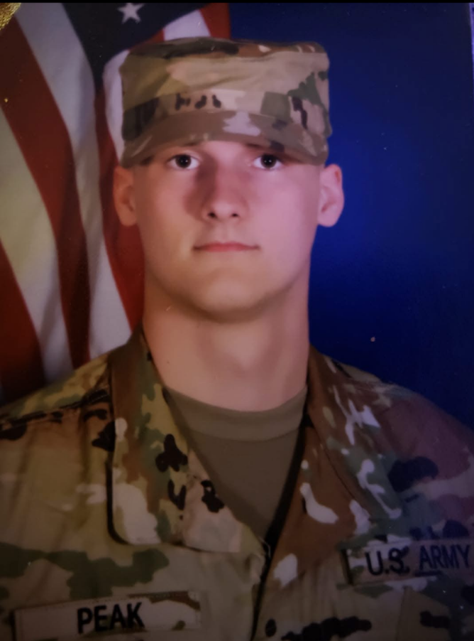 Young soldier in camouflage uniform standing in front of an American flag backdrop.