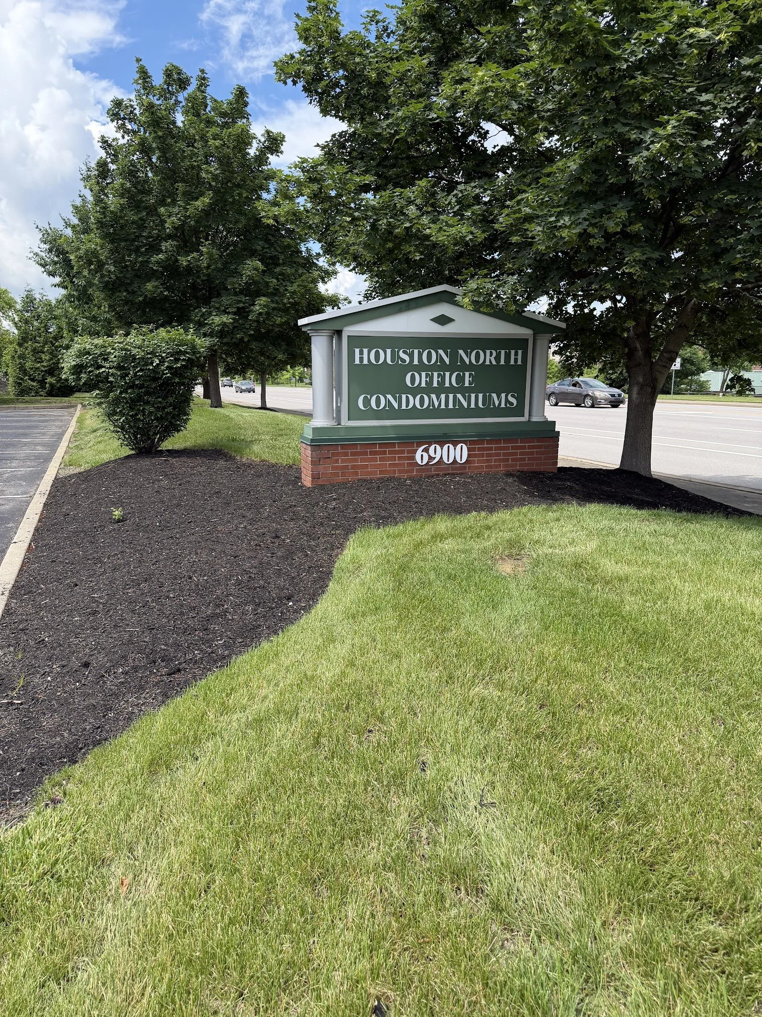 A sign for Houston North Office Condominiums at 6900, situated on a landscaped area with grass and bushes, during daytime with a partly cloudy sky. This is the sign you will see when entering the parking lot of The Purple Wax Suite in Florence, KY.