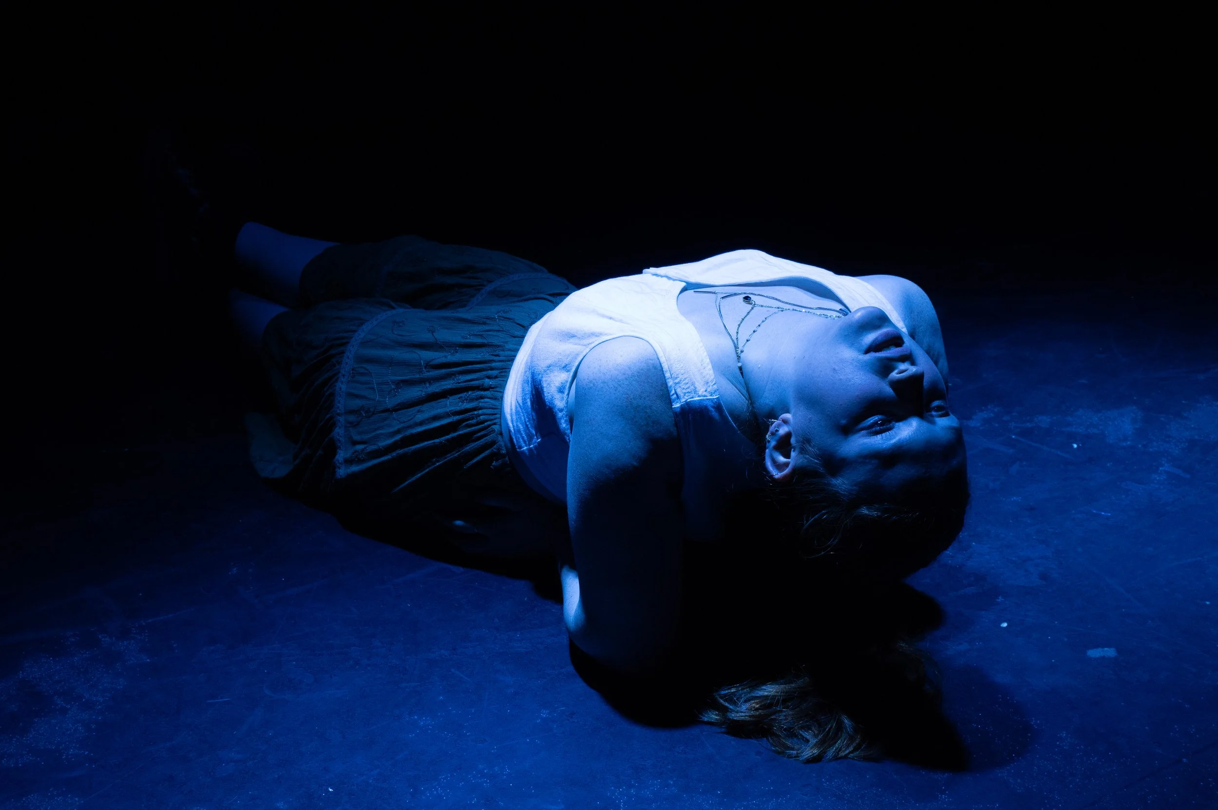 A woman lies on the floor in a dark room, illuminated by a blue light, with her eyes closed and her hands behind her head.