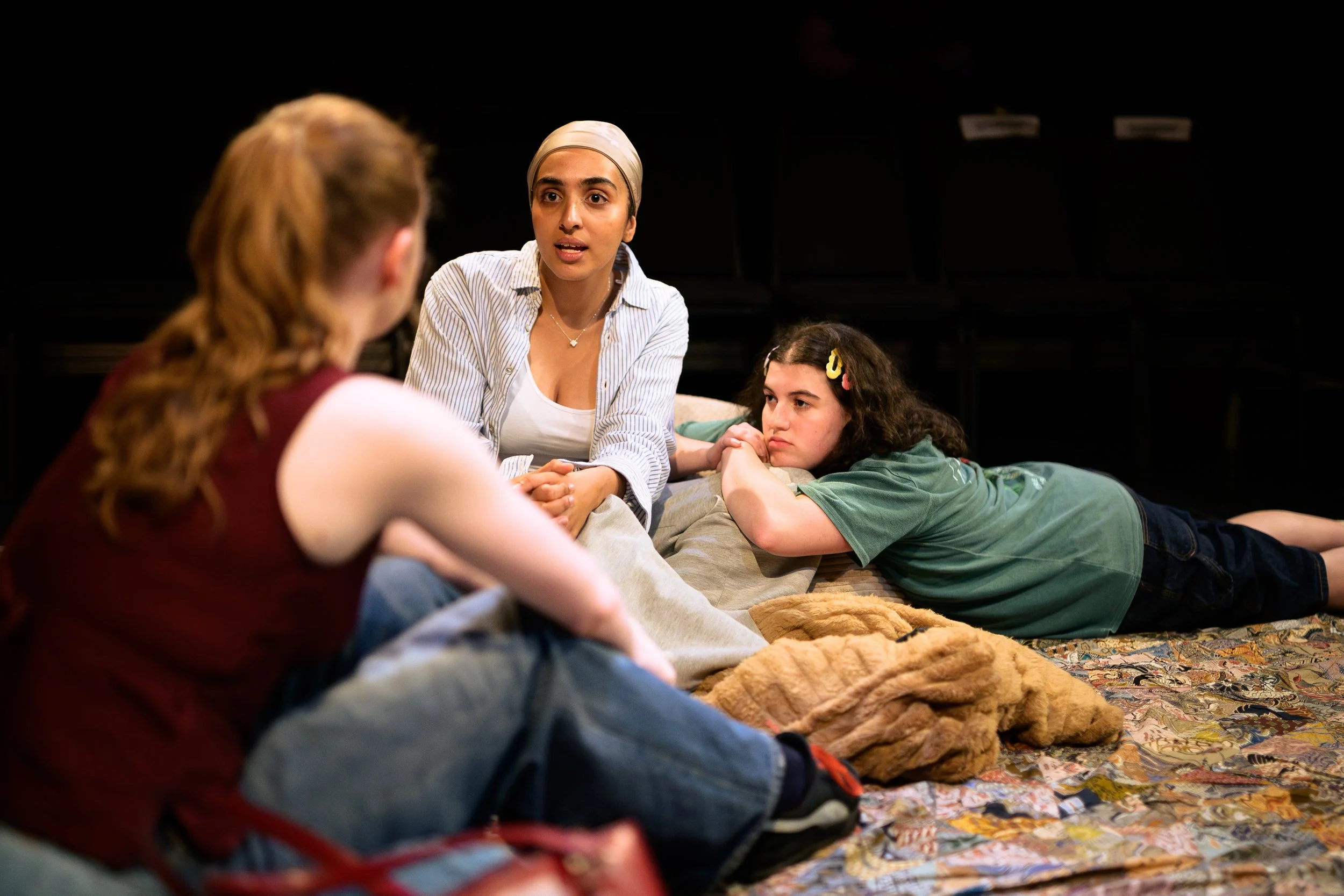 A woman wearing a beige head covering and striped shirt talking to two children, one with red hair in a braid and the other with dark curly hair lying on their stomachs on a colorful quilt, in a dark room with foldable chairs in the background.