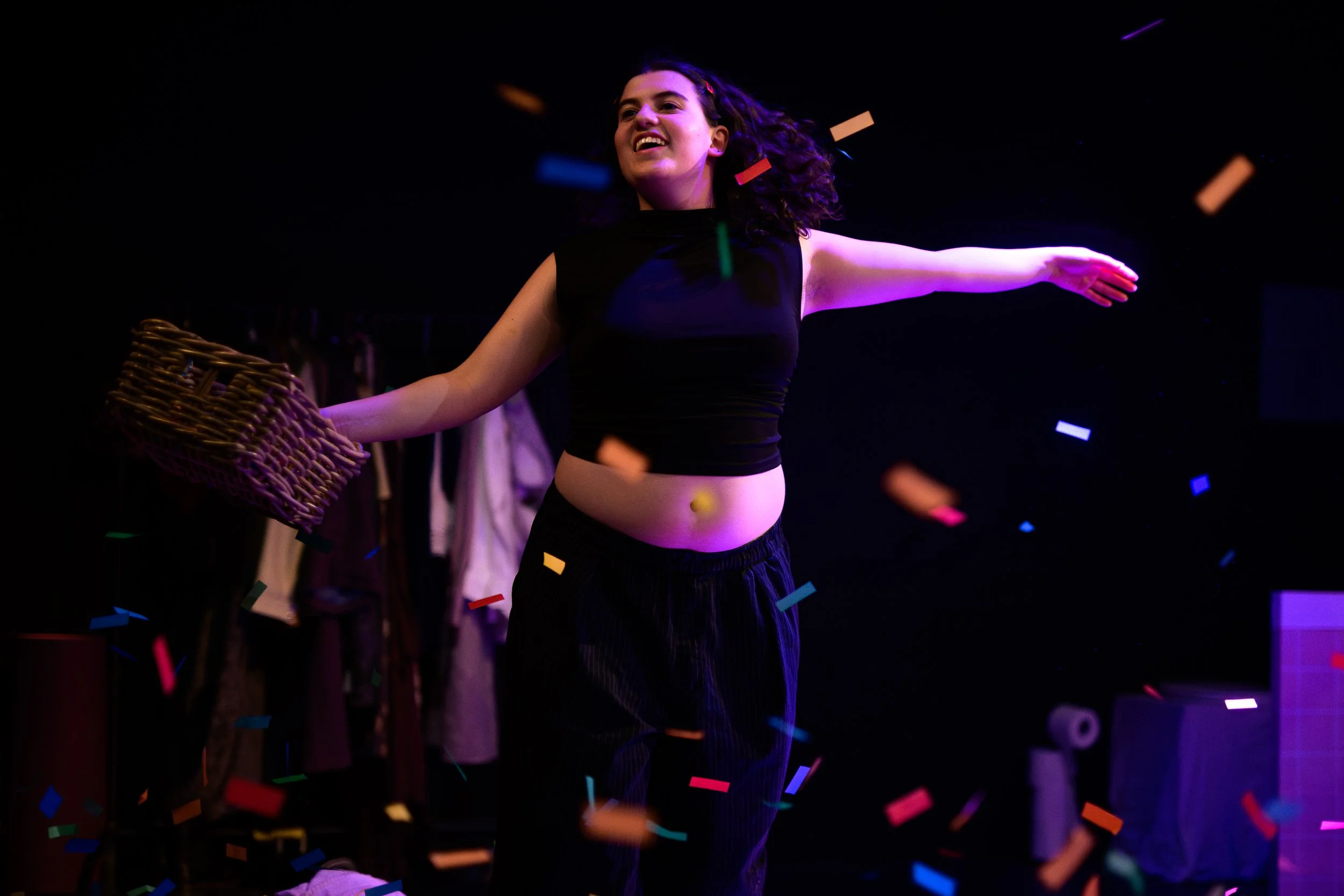 A young woman with curly hair smiling and dancing among colorful confetti in a dark setting with purple lighting.