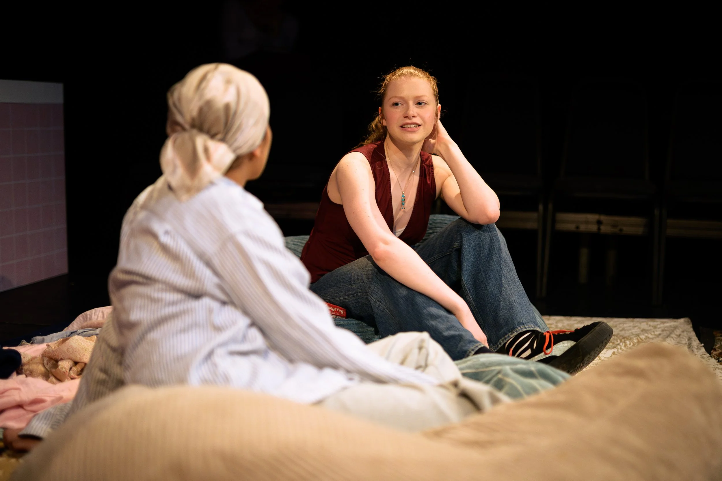 Two young women sitting on the floor, engaging in conversation. One has blonde hair tied back, wearing a white striped shirt, and is facing away. The other has red hair, wearing a sleeveless maroon top and jeans, and is facing the camera.