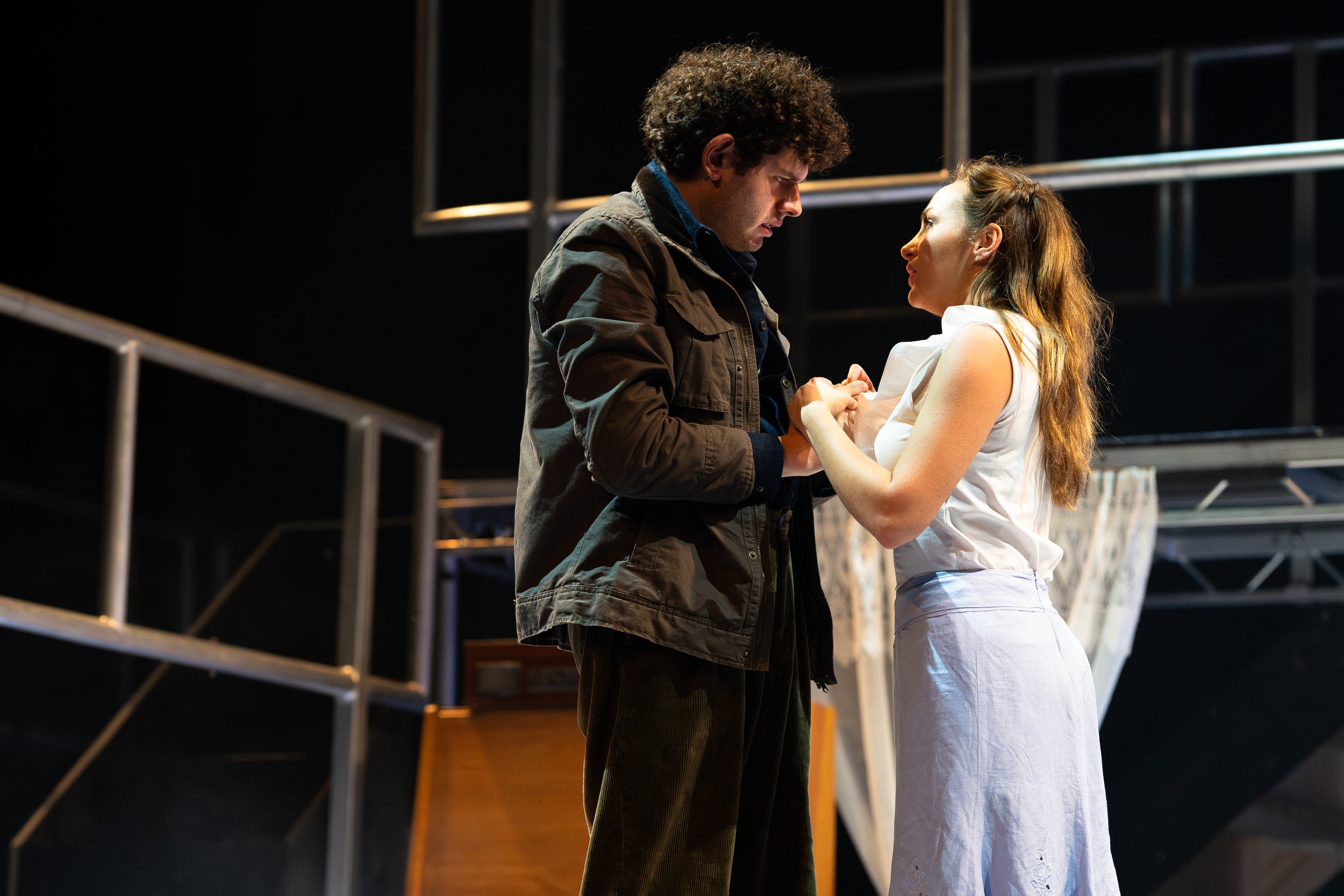 A man and woman are standing close, holding hands, looking into each other's eyes on a stage or theater set with metal railings.