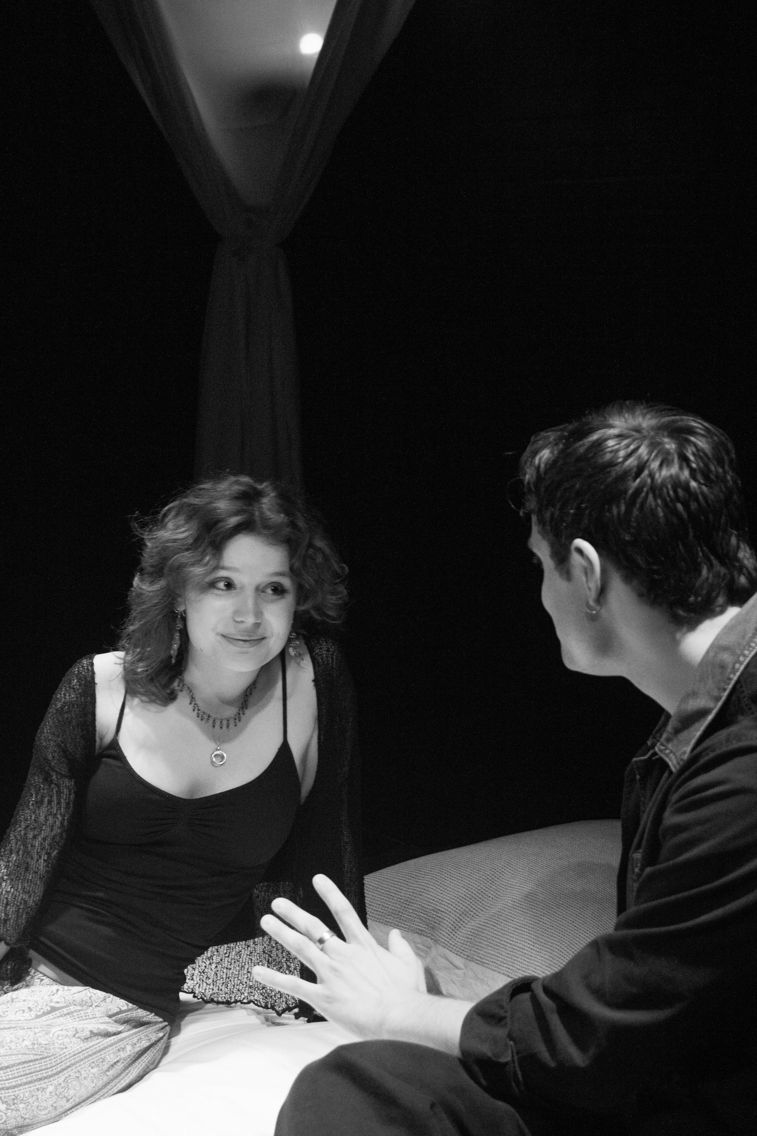Two women and one man engaged in a conversation in a dimly lit room. The woman on the left has curly hair, wears a black dress with lace sleeves, and is smiling. The woman on the right, with short hair, is seen from the side, wearing a dark jacket. T