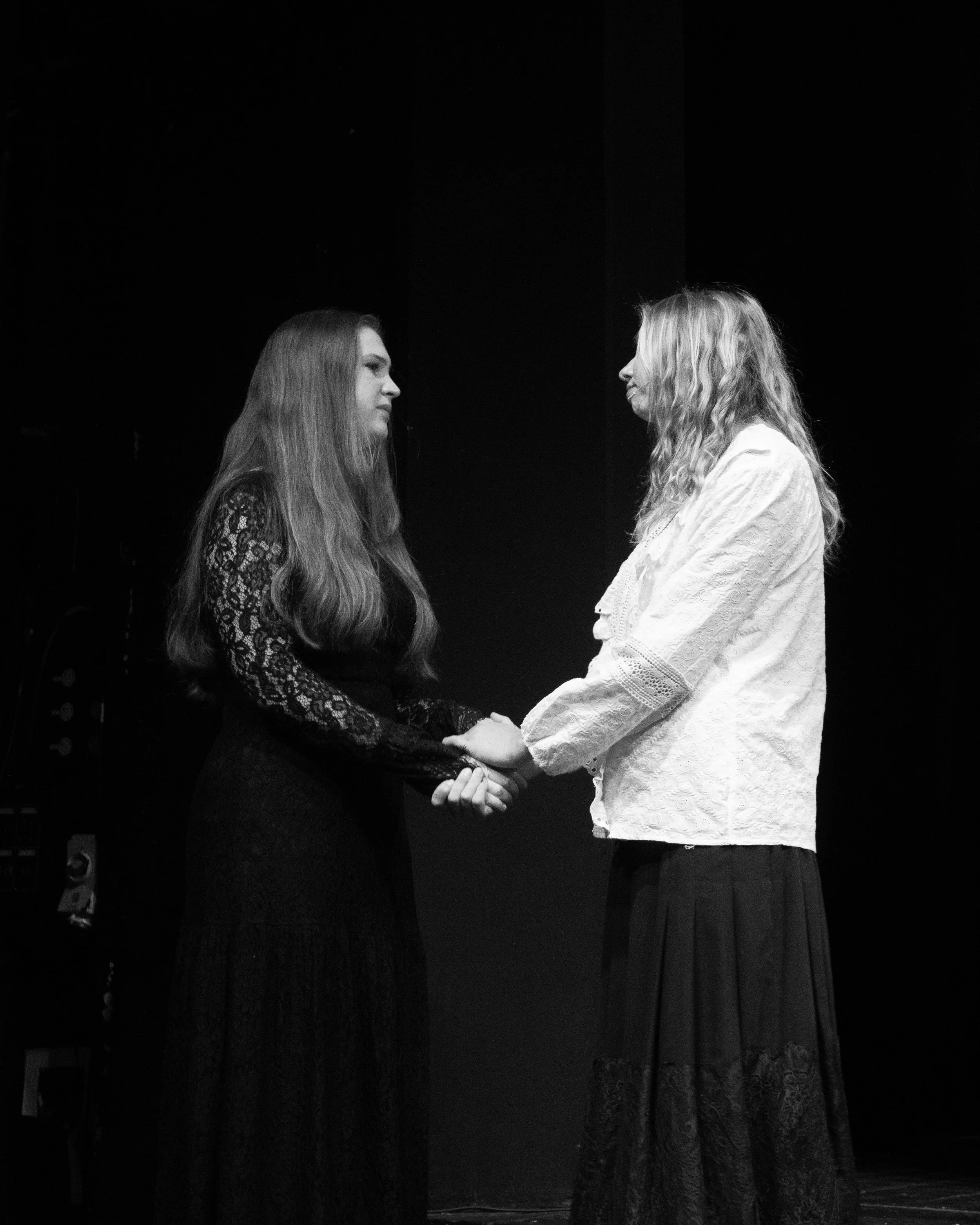 Two women are standing face to face, holding hands, on stage with a dark background. One woman is wearing a long dark lace dress, while the other is wearing a white lace dress. They are smiling at each other.