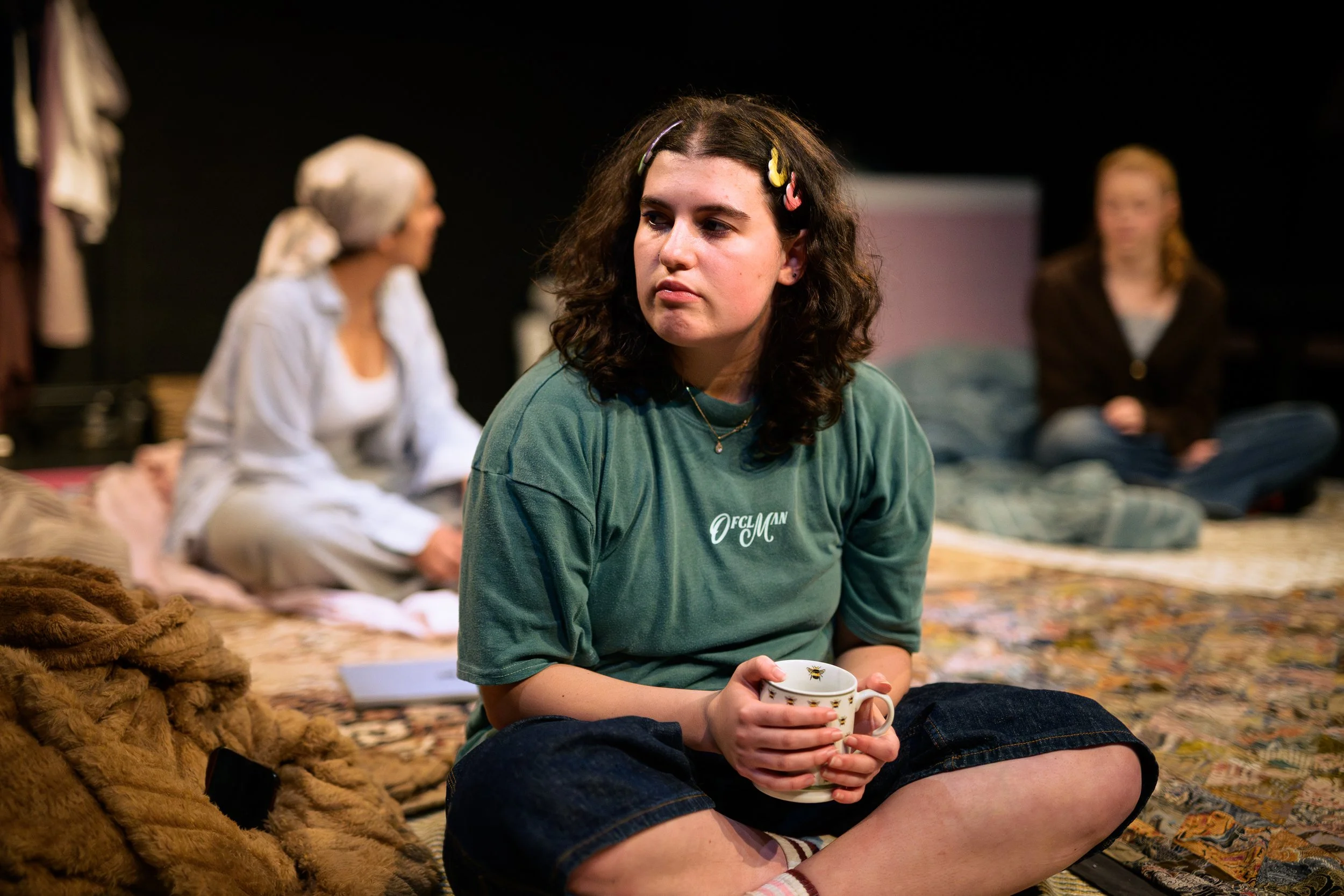 A young woman sitting cross-legged on a colorful rug, holding a coffee mug with a bee design, with other women sitting in the background, in a cozy indoor setting.