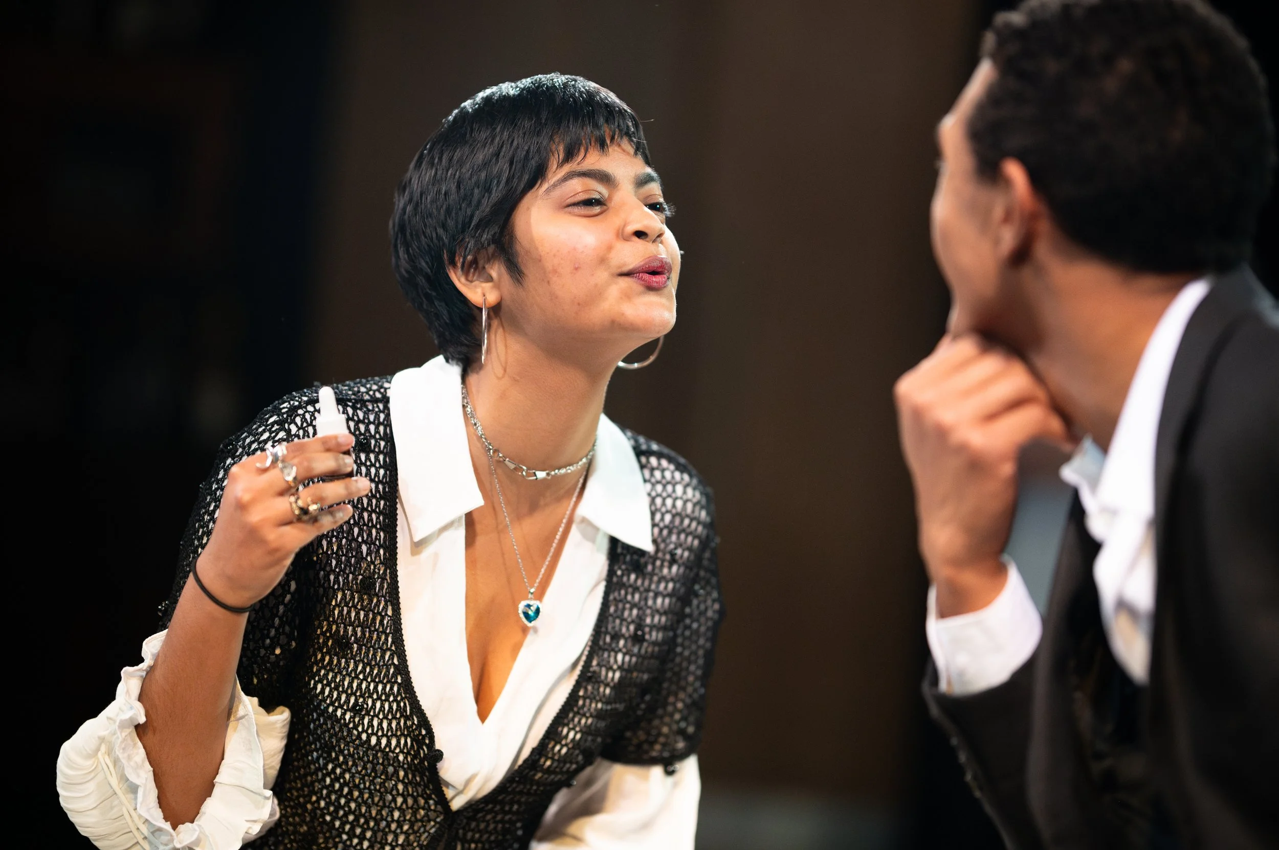 A woman with short dark hair and earrings is engaged in conversation with a man whose face is not visible. She is wearing a black and white outfit with jewelry, and is speaking animatedly.