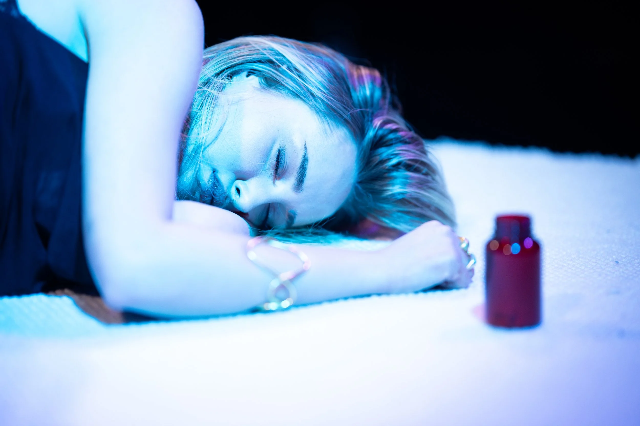 A woman with colorful hair lying on her side with her eyes closed, resting her head on her arm, in a dimly lit setting with blue lighting. There are small bottles in the foreground.