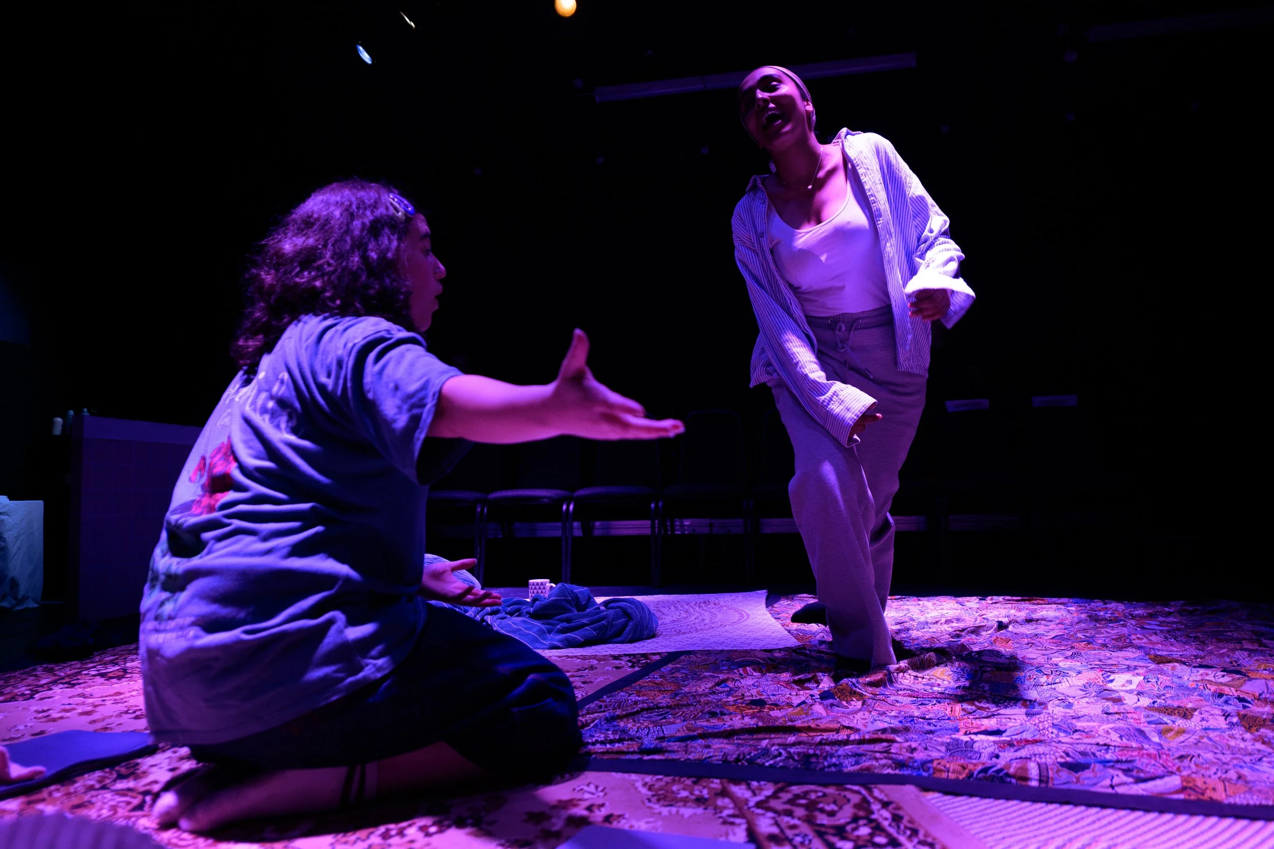 Two women are engaging in an intense conversation on stage, one is standing and smiling while the other is seated on the floor, appearing to be in thought. The scene is lit with purple stage lighting.