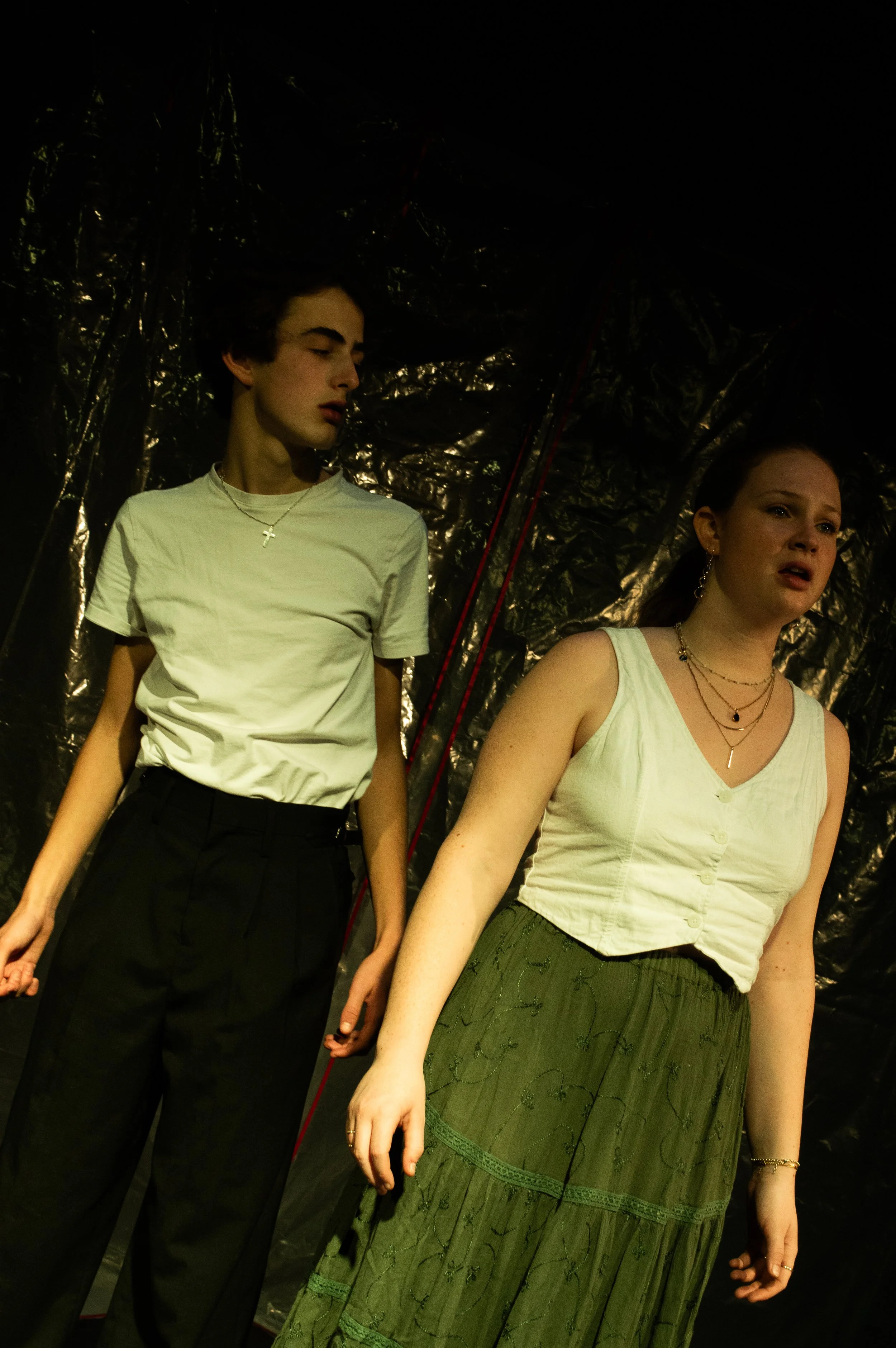 Two young women holding hands, standing in front of a shiny, crinkled metallic background. One woman wears a white tank top and a green skirt, while the other wears a white t-shirt and black pants. They are both wearing jewelry, and the woman on the 