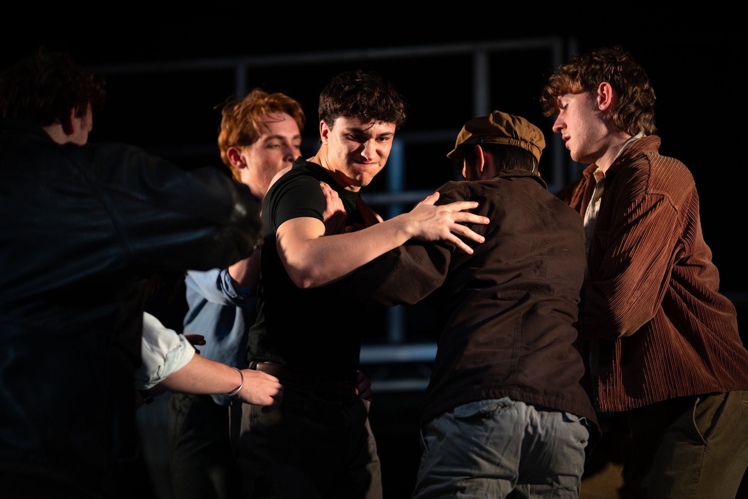 Group of young men, some with curly hair, engaged in an intense conversation or fight outdoors at night, with a dark background.