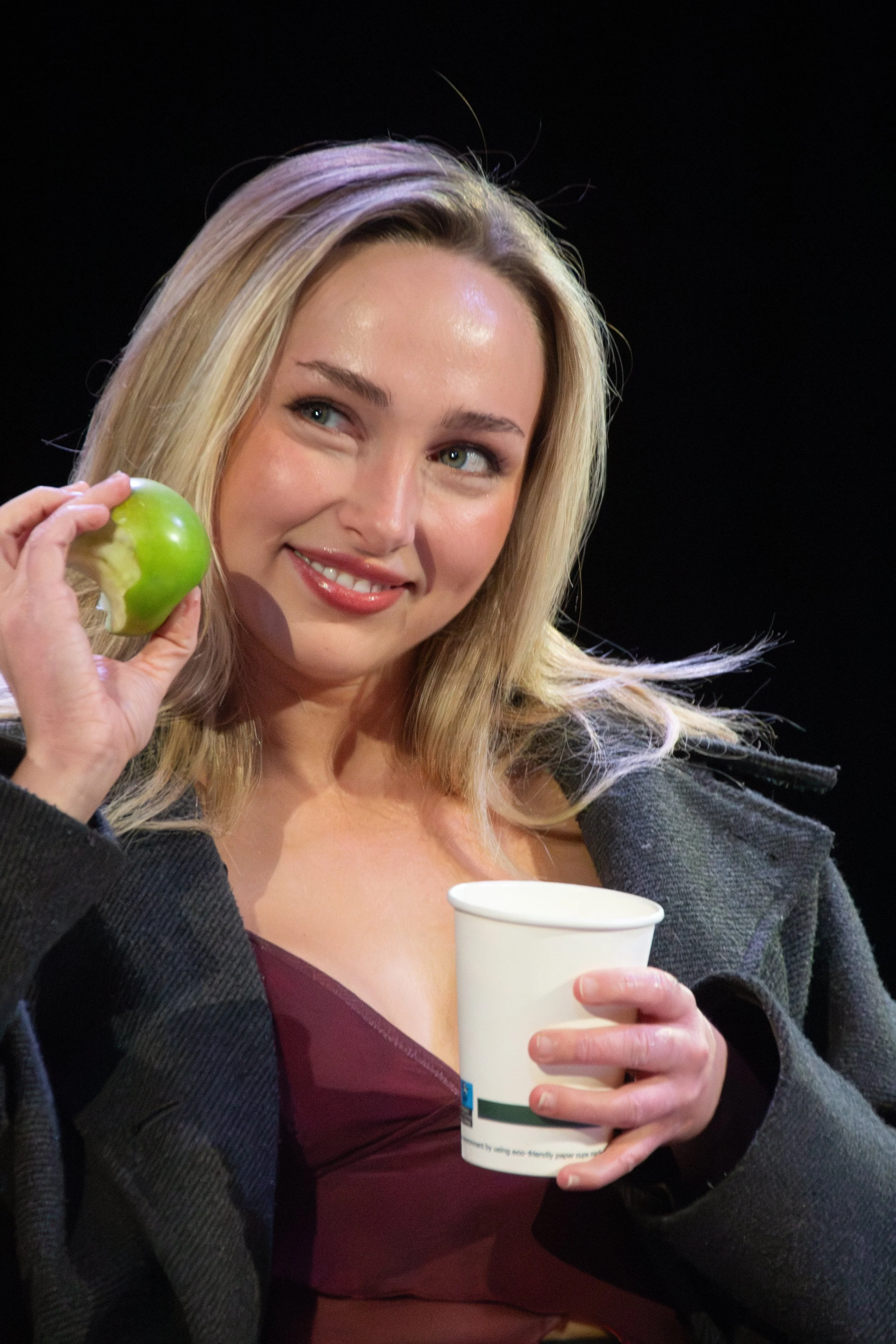 A woman with blonde hair and a warm smile holding a green apple near her face, wearing a dark blazer and a burgundy top, and holding a white paper cup in her other hand.