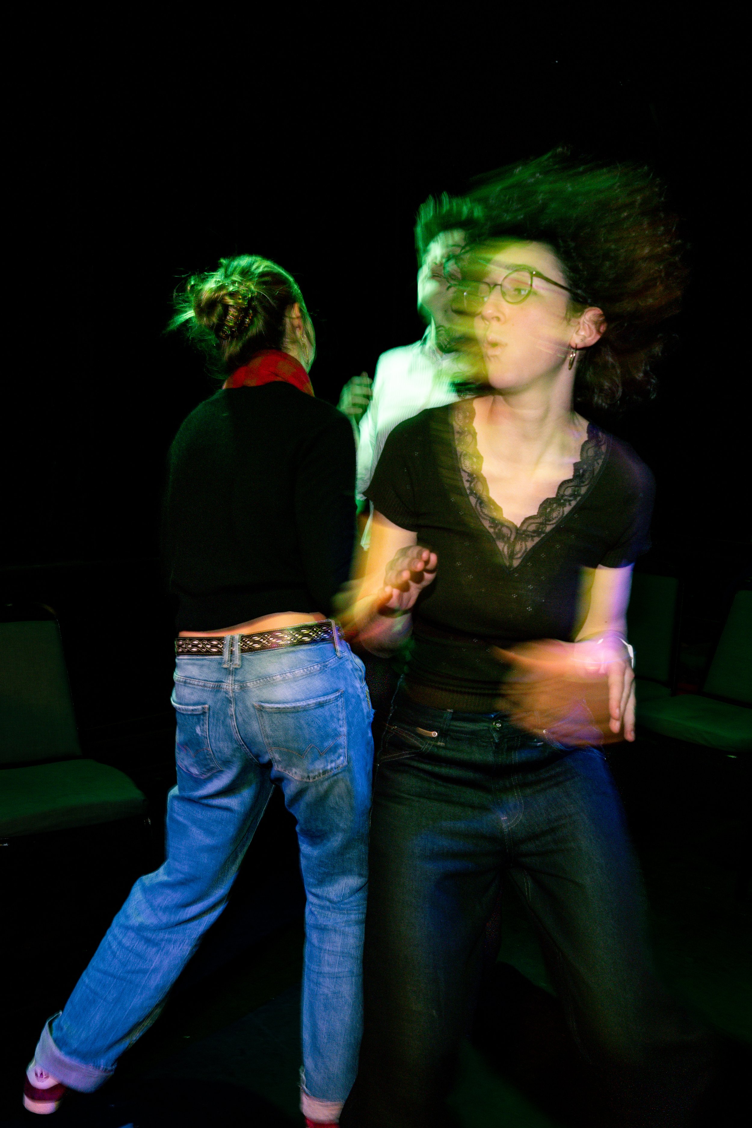 Two women dancing at night, one wearing glasses and black shirt with lace, the other with a bun hairstyle, jeans, and a black blazer. Blond hair in motion.