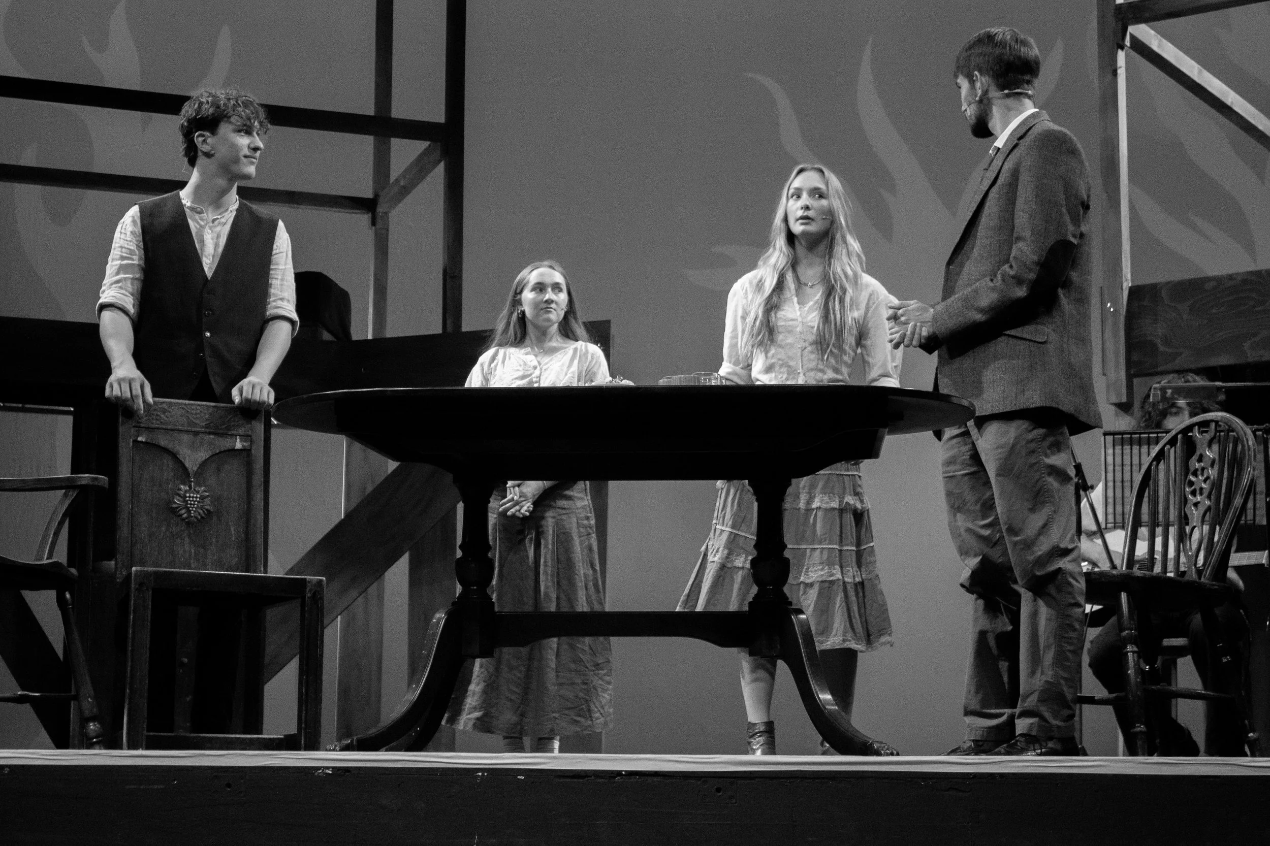Black and white photo of four young actors performing on stage. One male actor stands behind a wooden podium to the left, looking at a female actor dressed in a long skirt and blouse in the center. Another female actor stands behind a round table, lo