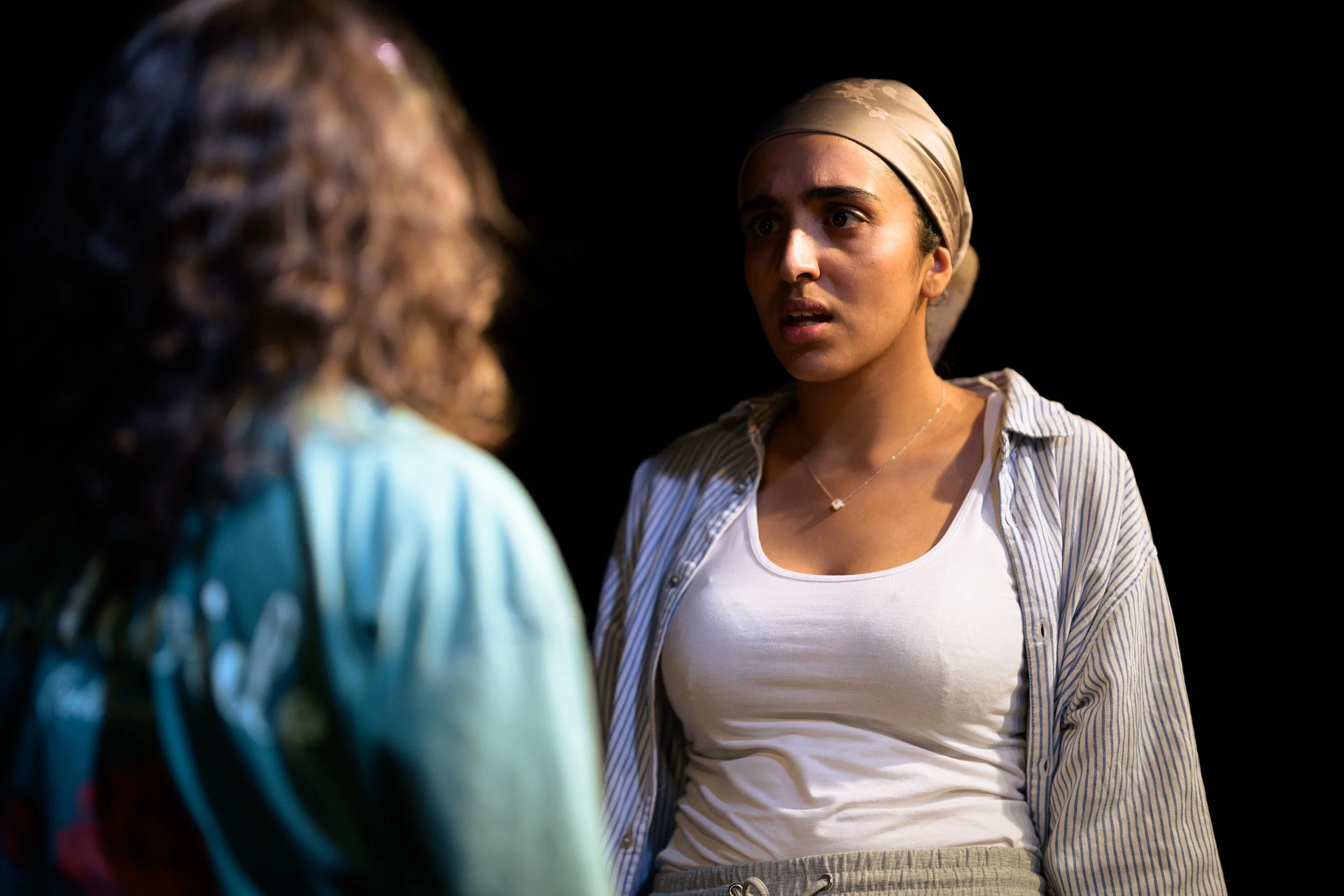 Two women engaged in conversation, one with curly hair and colorful clothing, the other wearing a beige headscarf, a striped shirt, and a white top, against a black background.