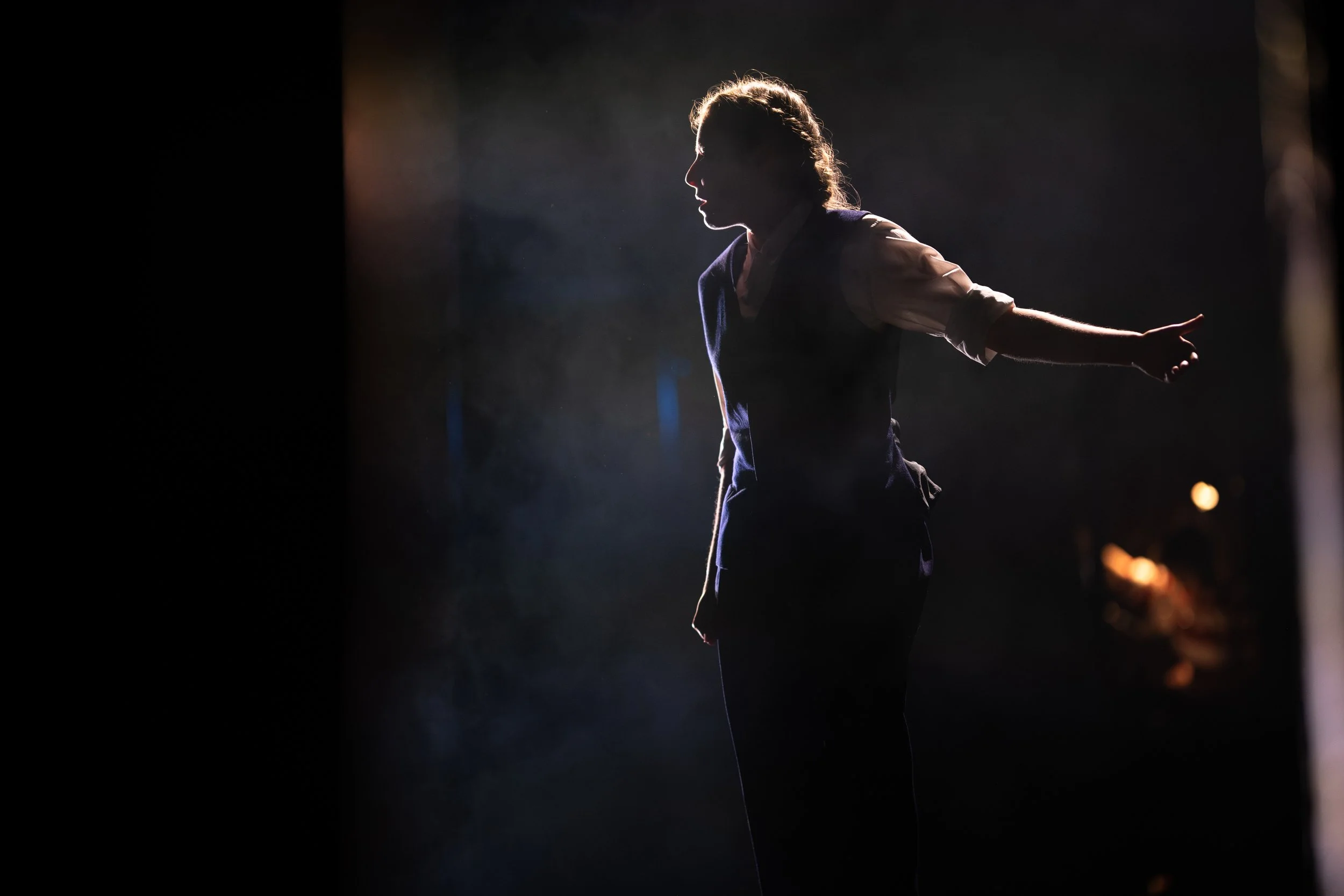 A silhouette of a woman with outstretched arms standing in front of a dark background with light illuminating her profile.