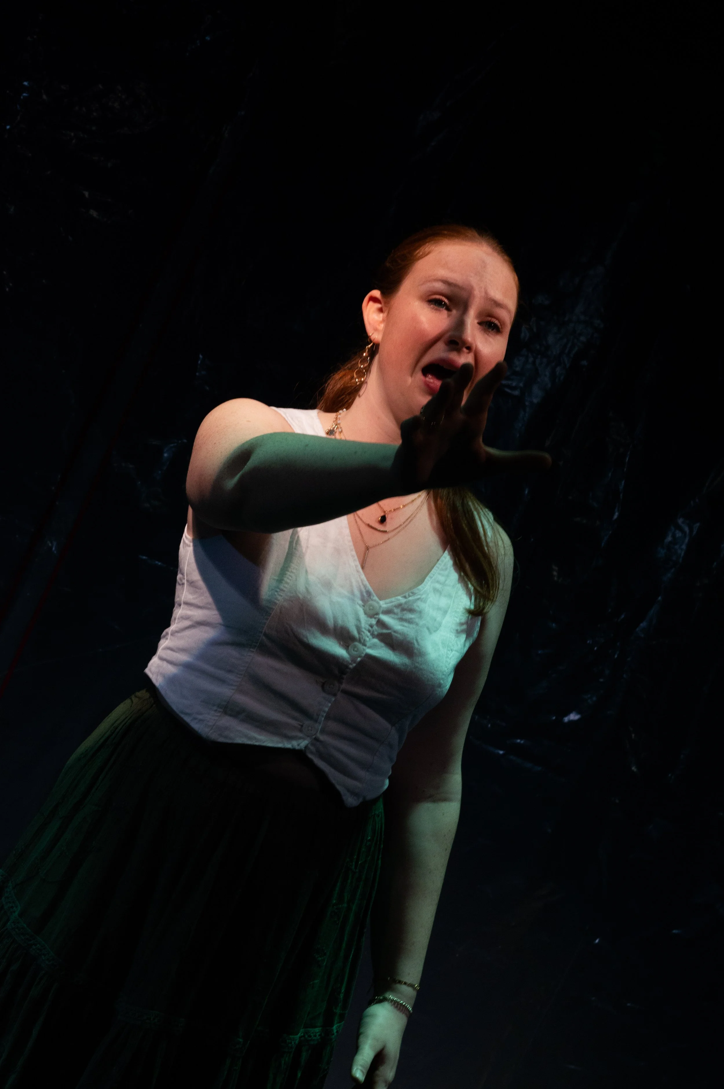 A woman with red hair wearing a white sleeveless top and a green skirt, reaching out with her right hand, in a dark setting with textured black background.