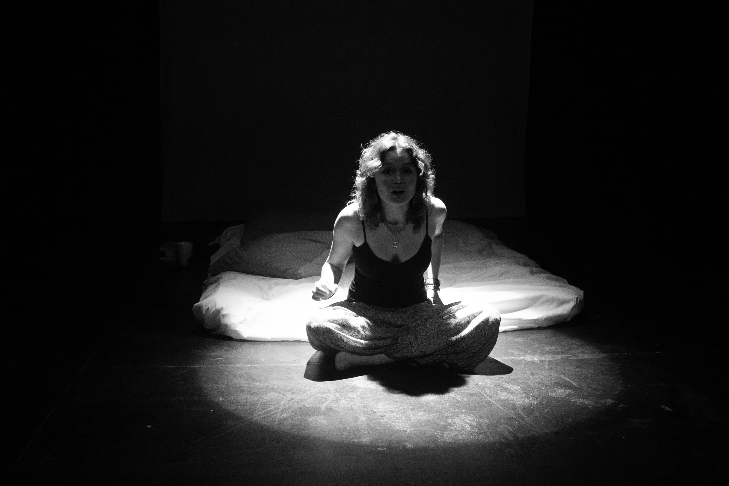 Black and white photo of a woman with curly hair sitting cross-legged on a bed in a dark room, illuminated by a single light source, with plain walls and a pile of clothes or belongings at the head of the bed.