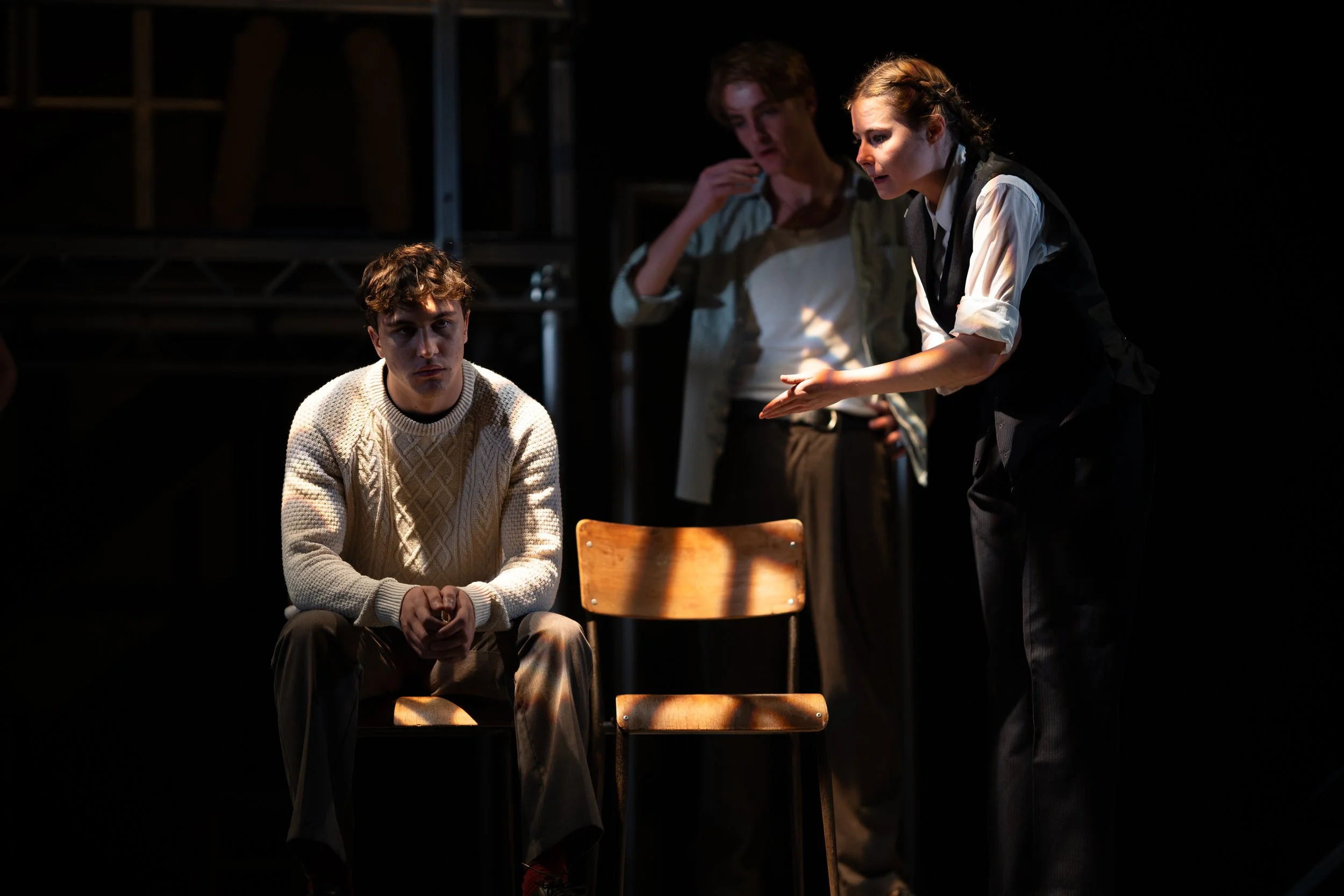 Three actors on a stage in a theatrical scene; a young man in a cream sweater seated in the foreground, a young woman with braided hair and a vest leaning towards him, and another woman taking notes in the background.