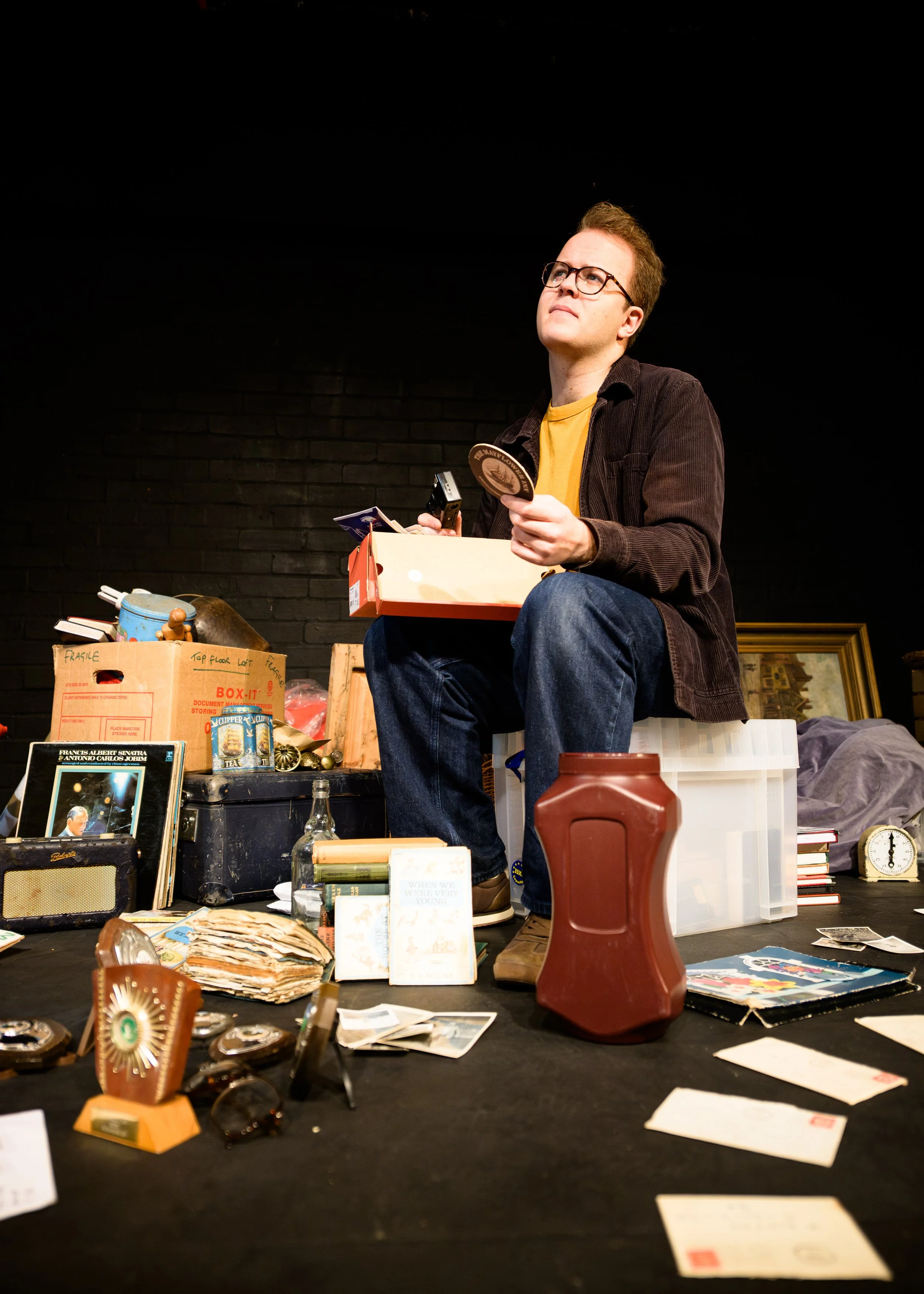 Person sitting on a white case, wearing glasses, holding a circular object, surrounded by various items like books, papers, and knickknacks on a table and boxes, with a dark background.