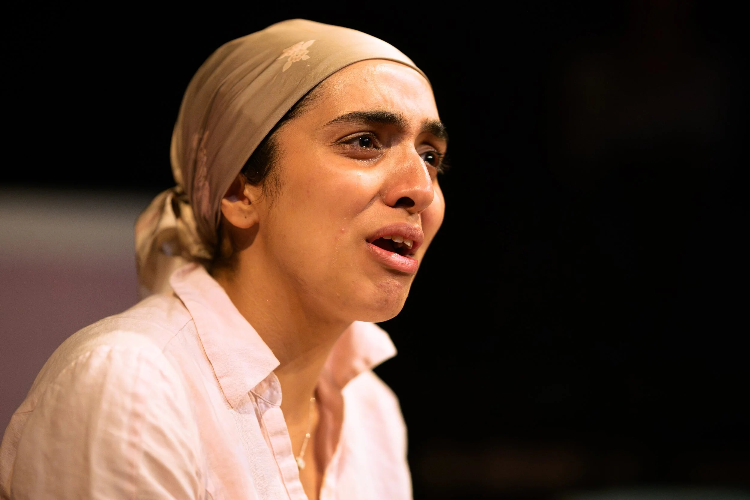 A young woman with dark hair, wearing a beige scarf and light-colored shirt, appears to be speaking or singing against a dark background.