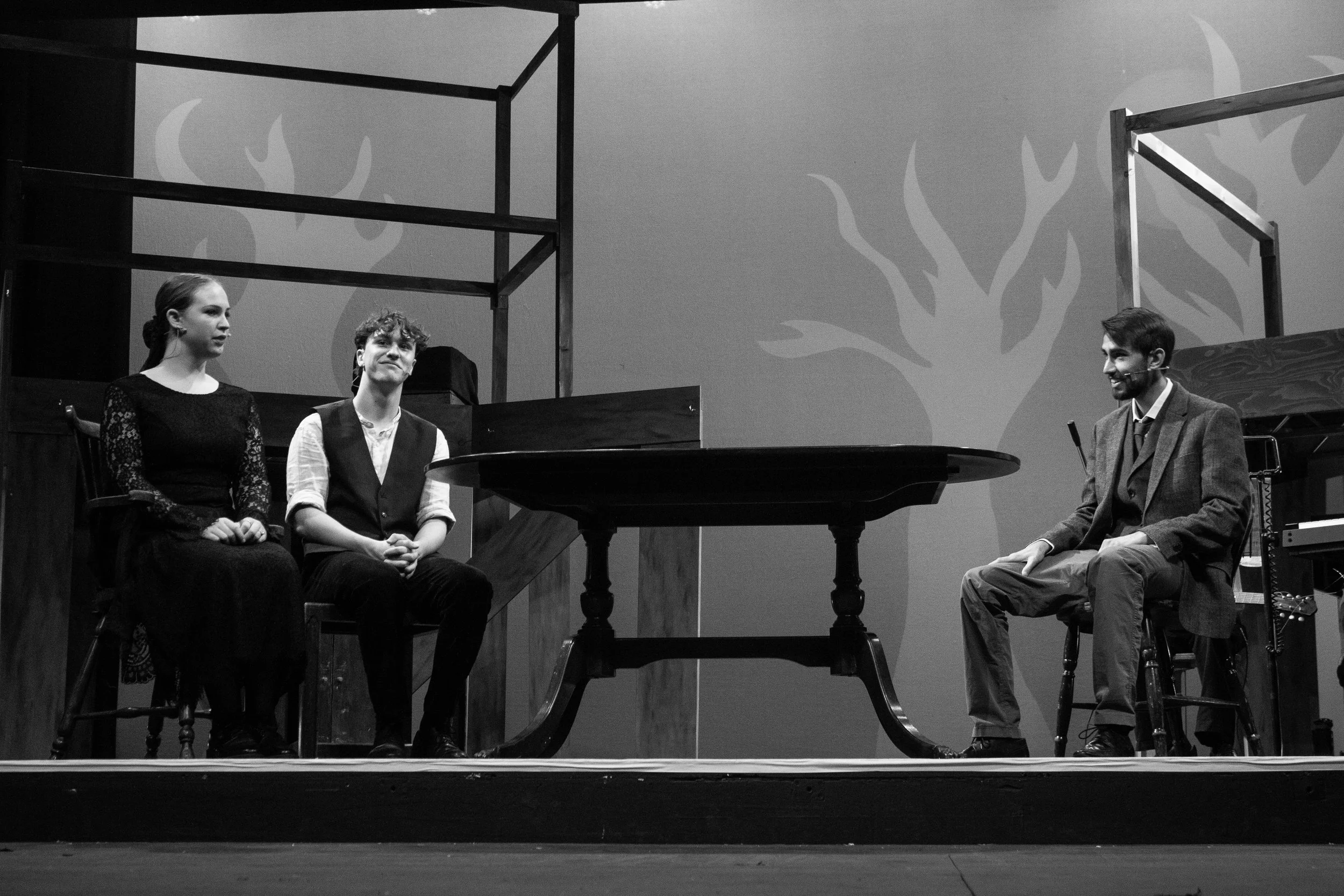 Black and white photo of three actors on a stage, two women and one man, engaged in a scene. The stage has a piano and simple wooden scaffolding. The background features a painted tree silhouette.