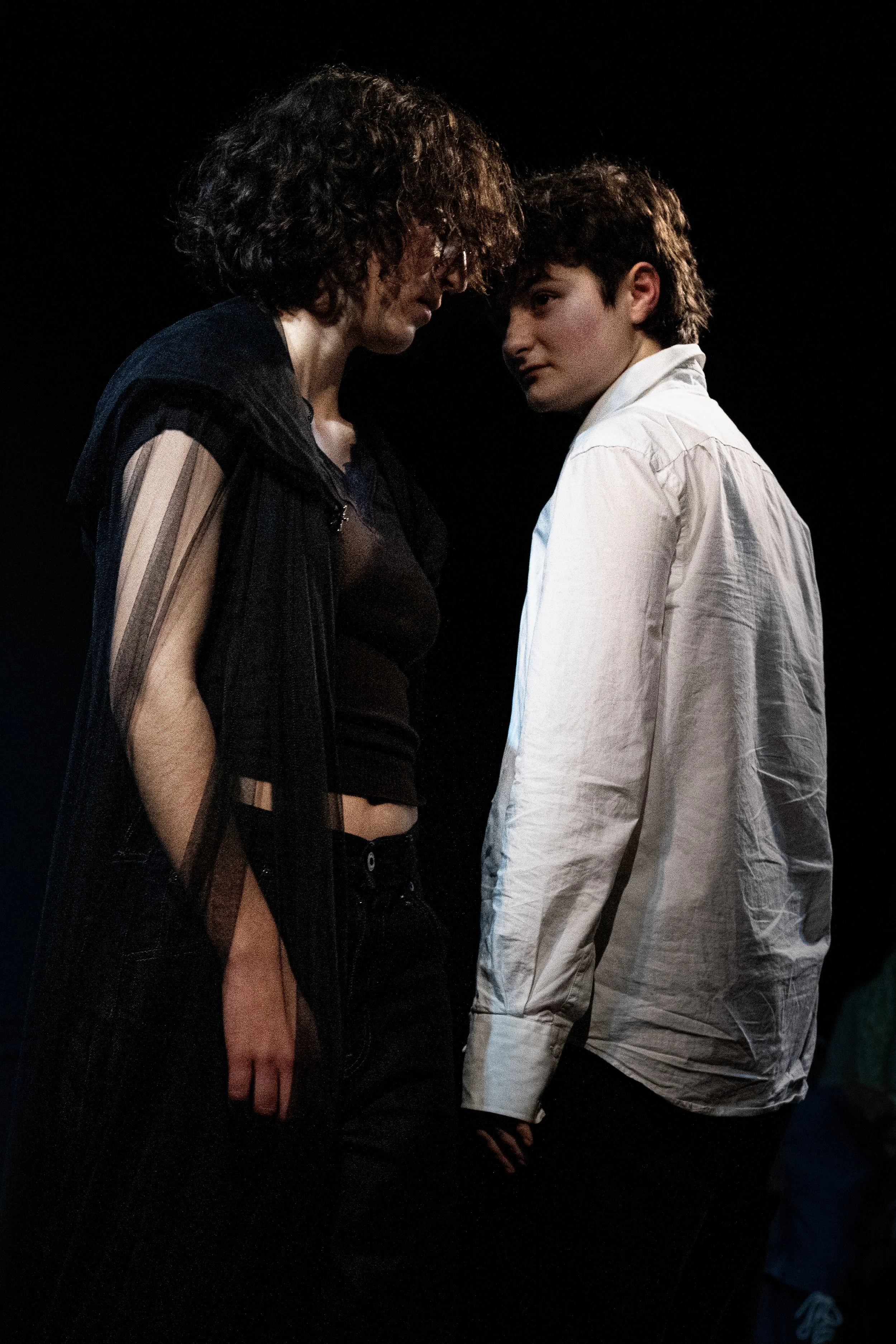 A woman and a young man are standing close with their foreheads touching against a dark background, creating an intimate moment.