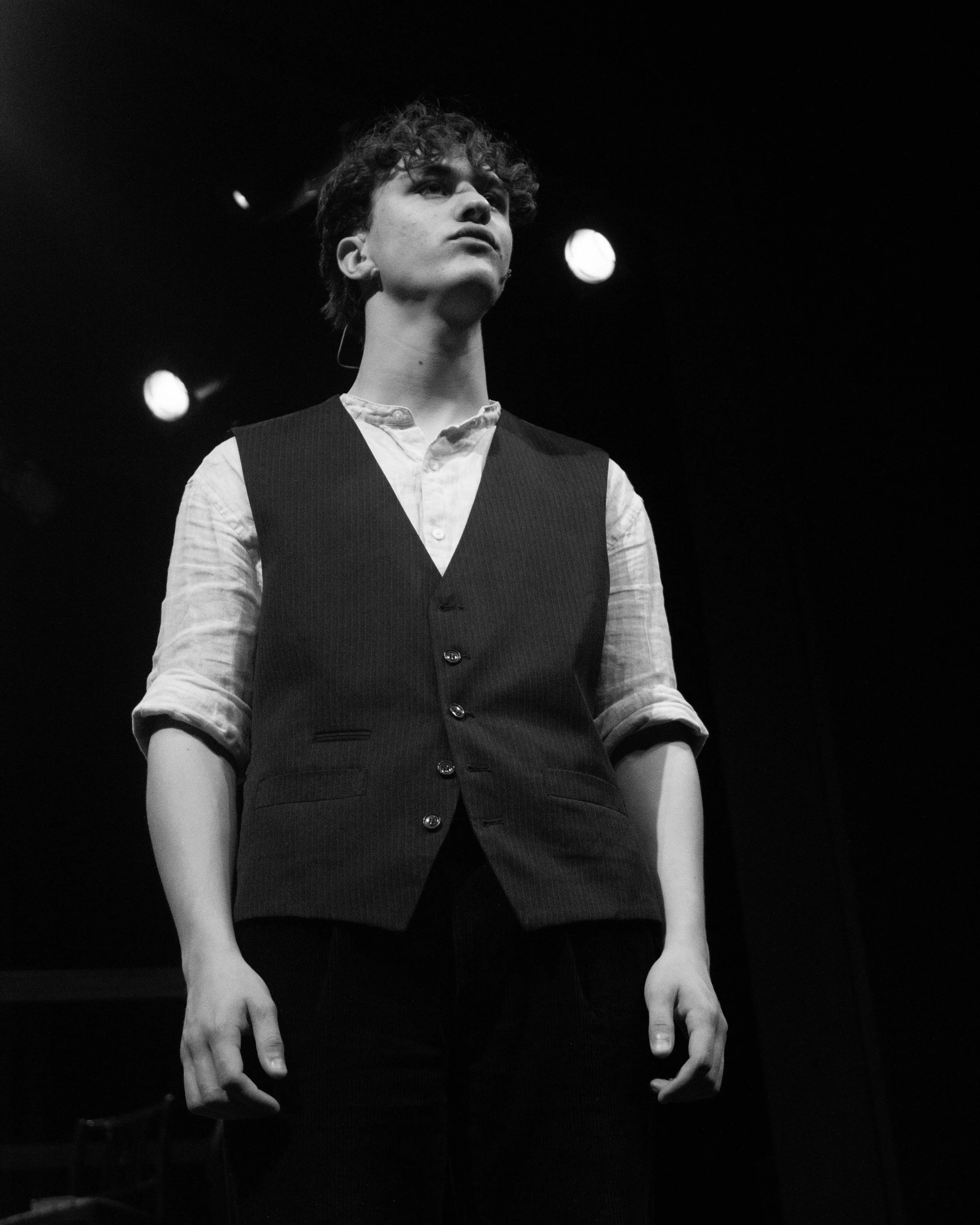 Black and white photo of a young person with curly hair, wearing a pinafore dress over a collared shirt, standing against a dark background with stage lights, looking serious and focused.
