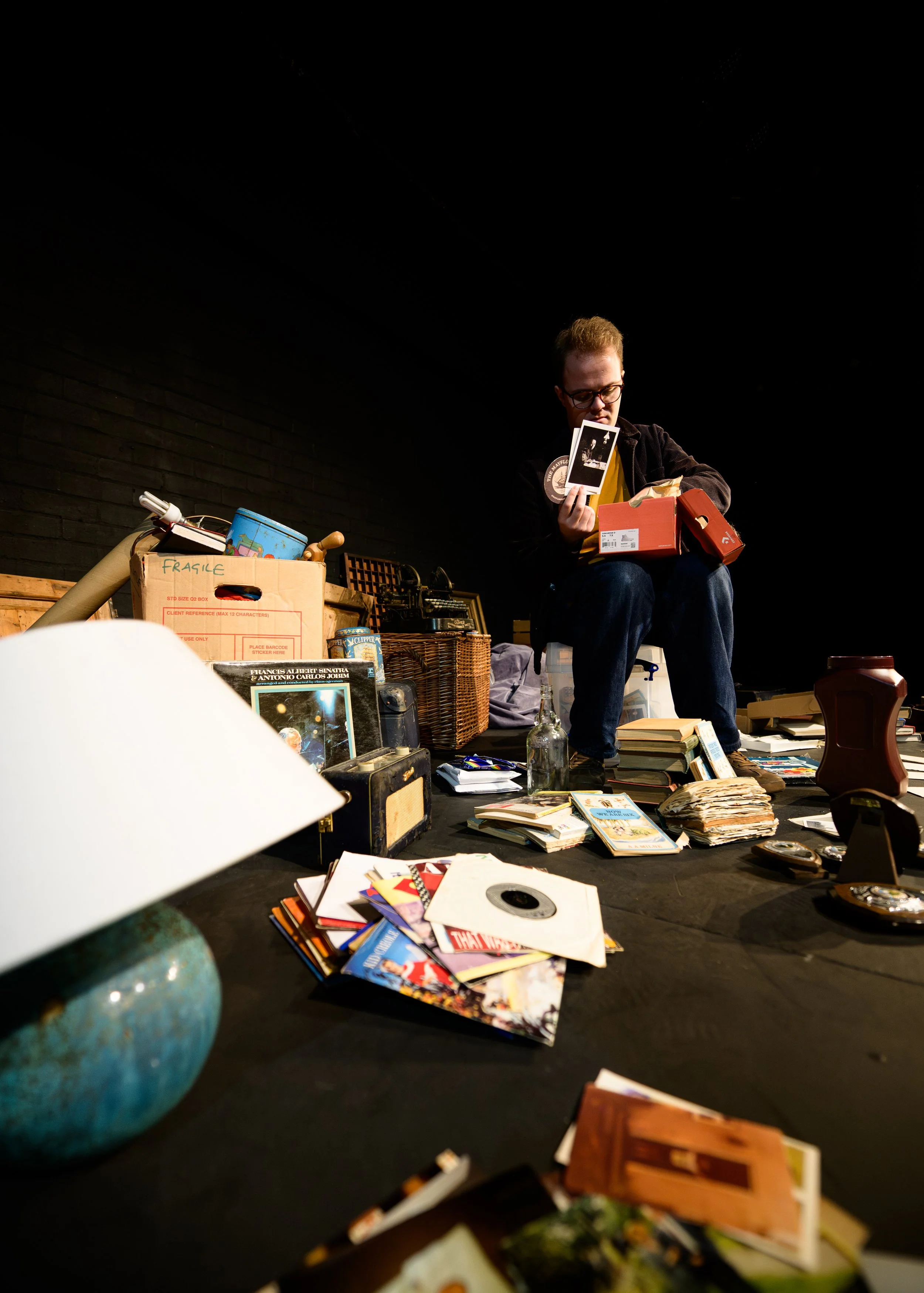 A man with glasses and a black jacket sitting on a chair surrounded by various books, papers, and boxes in a dimly lit room with a black brick wall.