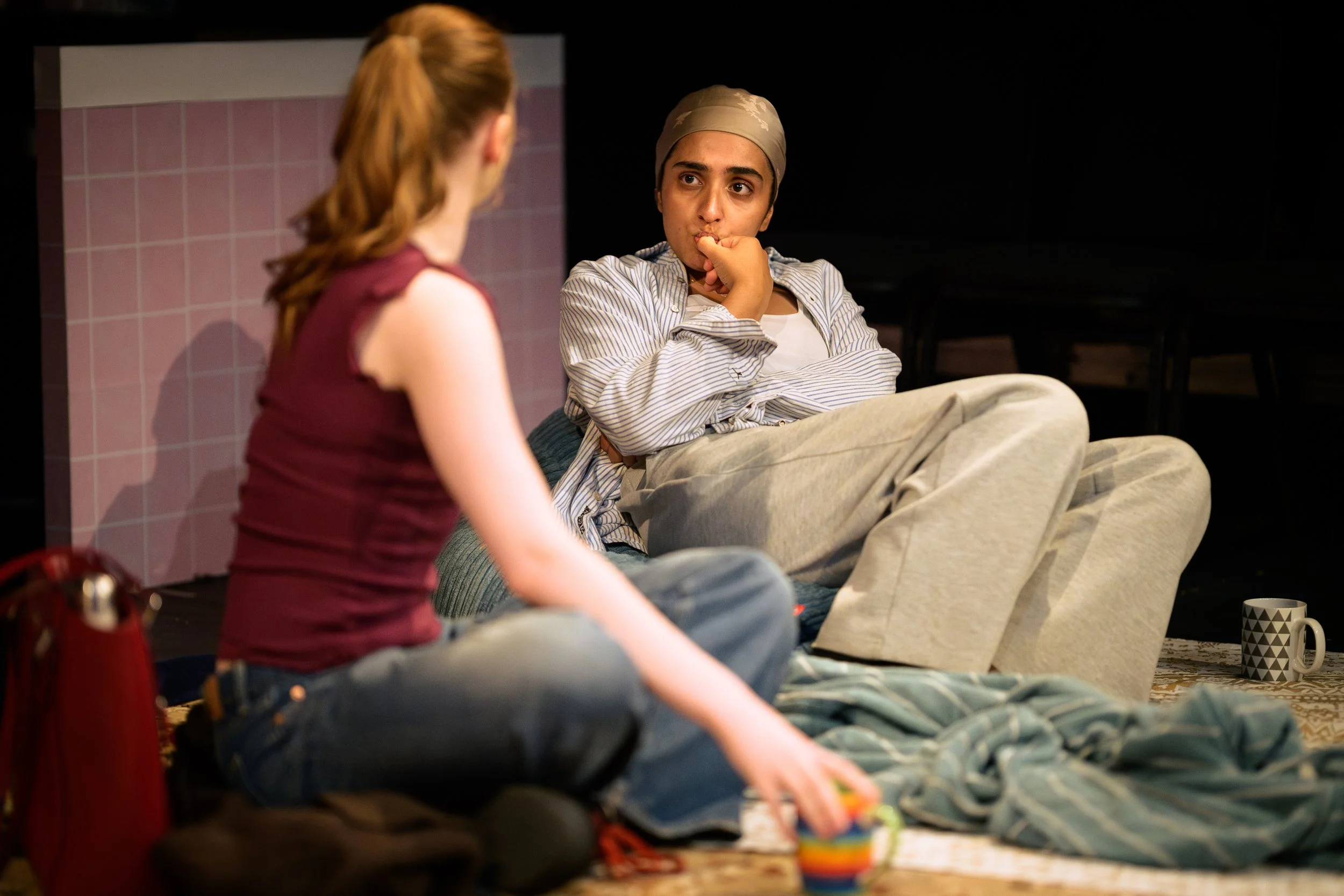 A person with a bandana on their head is sitting on the floor with crossed legs, looking at a woman with long red hair who is sitting nearby. There are cups and objects on the floor around them in a cozy indoor setting.