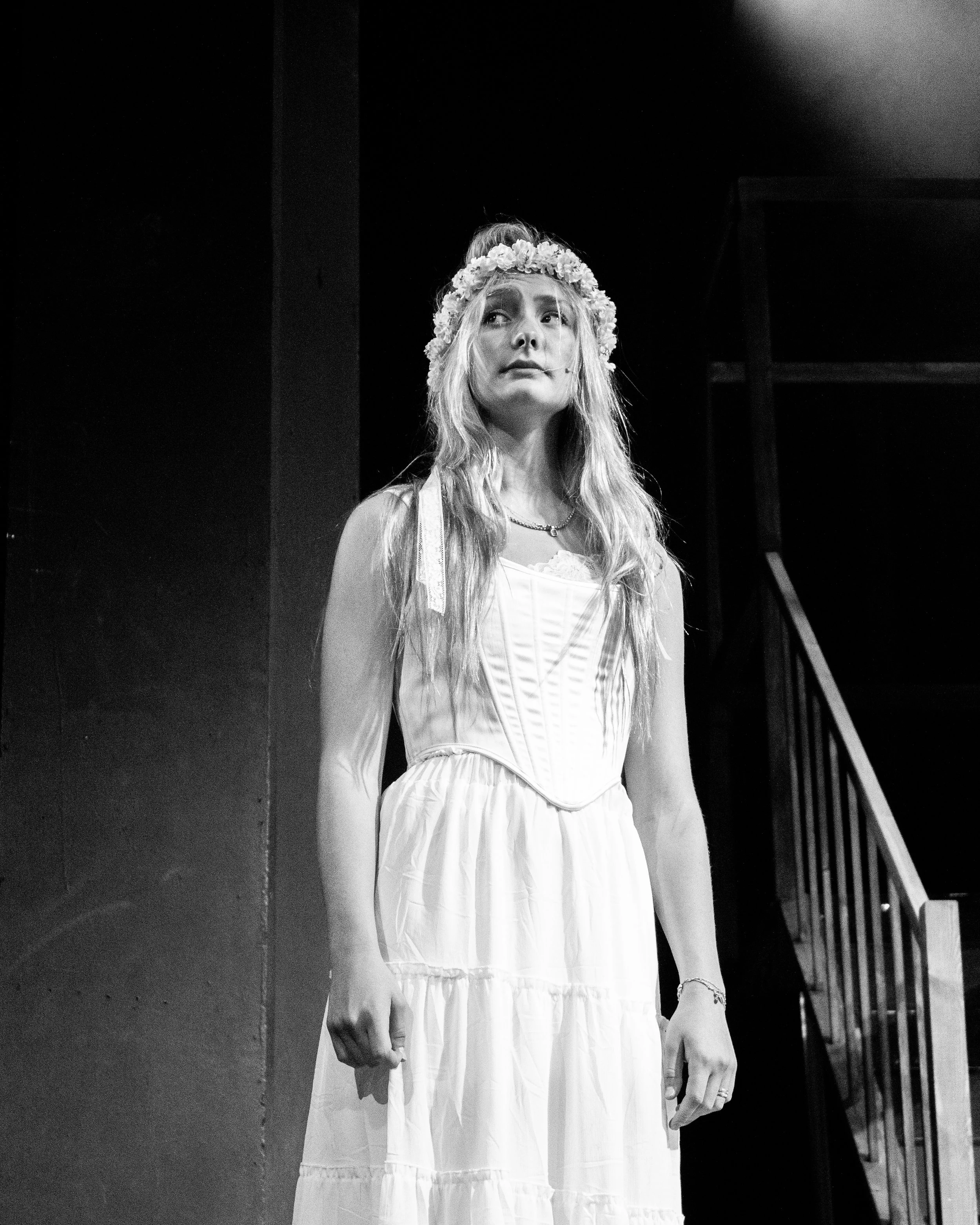 Black and white photo of a woman with long hair wearing a floral headband and a white dress, standing indoors.