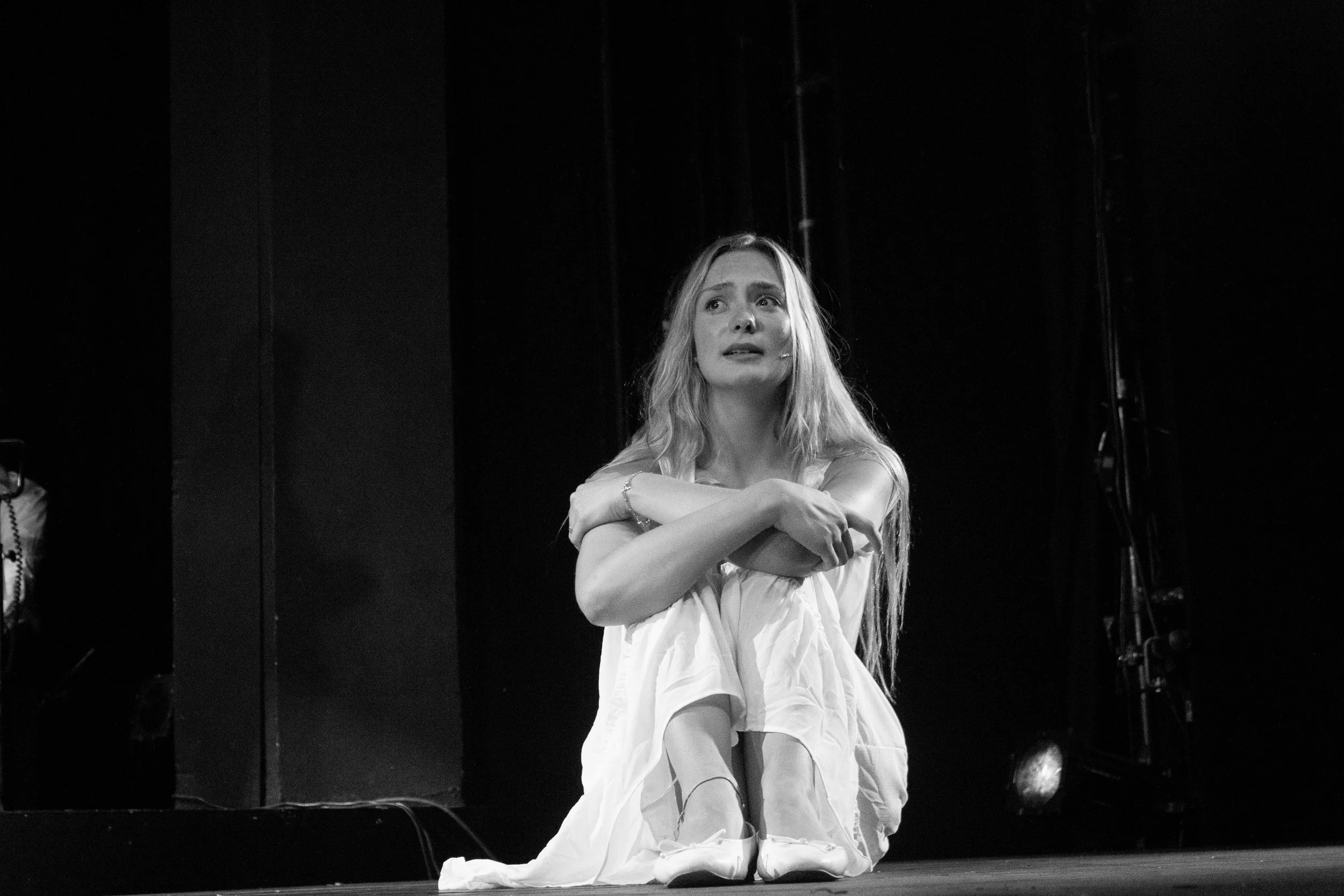 A woman with long, wavy hair sitting on the floor wearing a loose, white dress, with a distressed or thoughtful expression, in a dark room with a black background.
