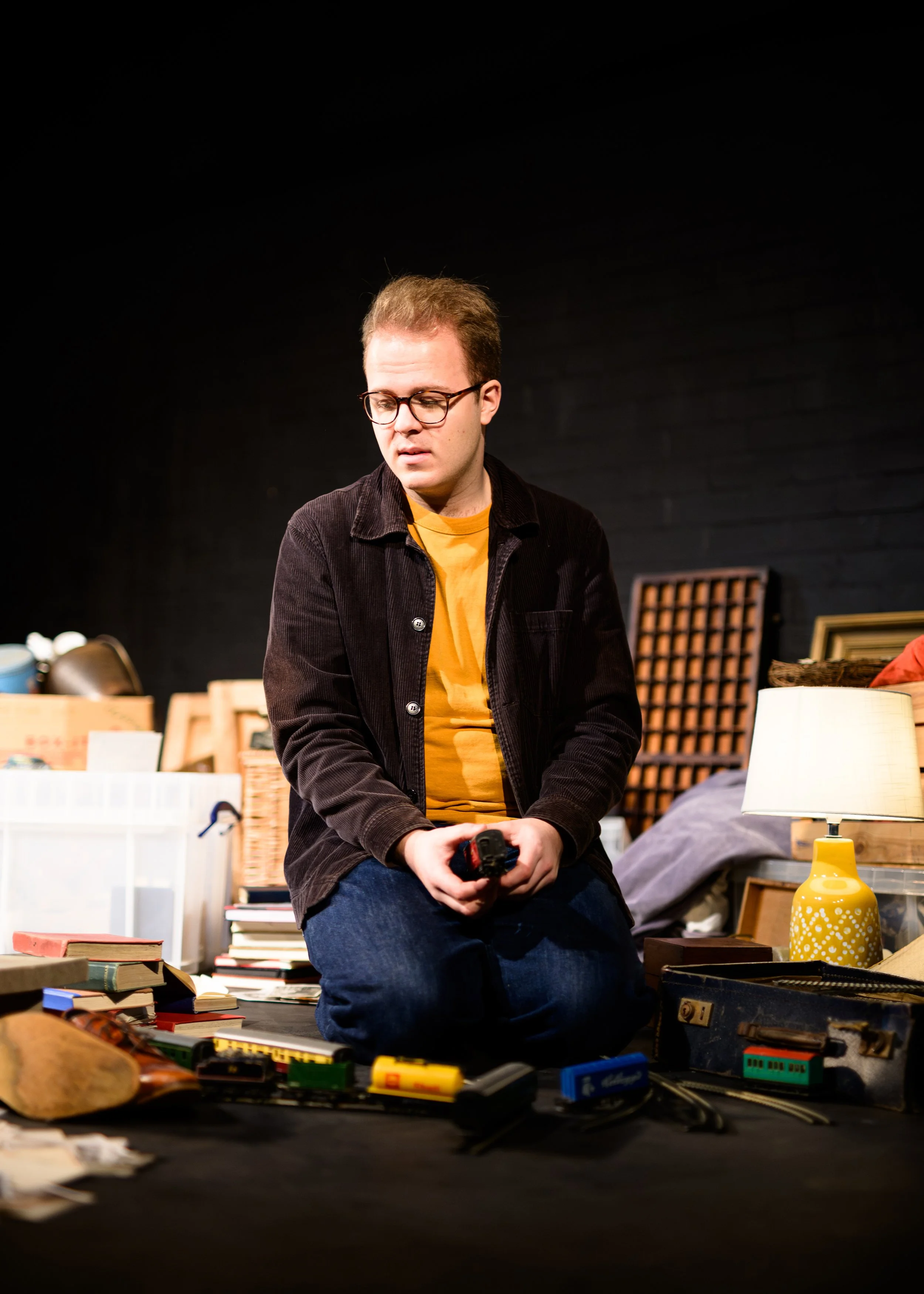 A young man with glasses sitting on the floor surrounded by books, toys, and various items, holding a remote control, with a black background and a yellow lamp nearby.