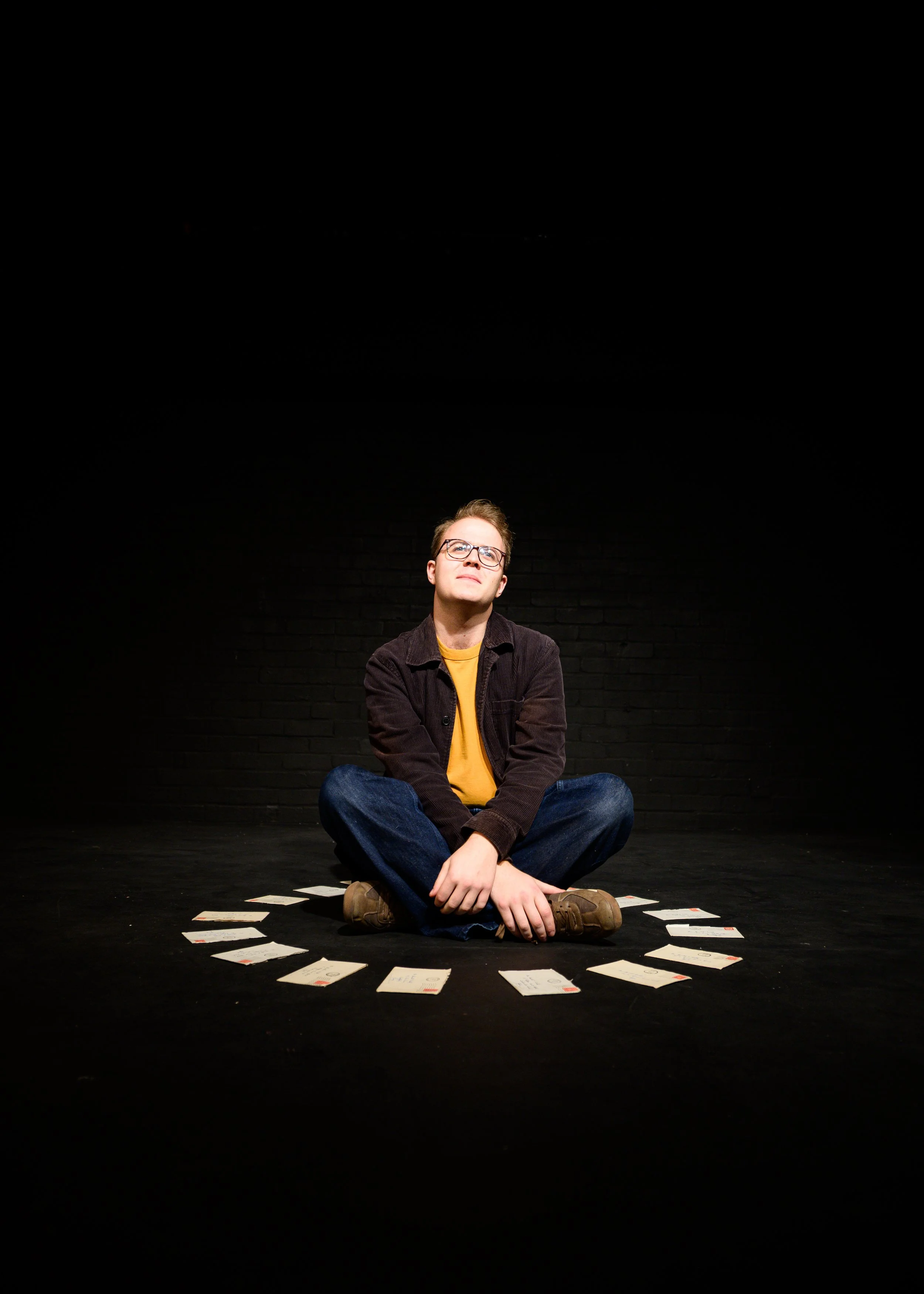 A young man with glasses, sitting cross-legged on a black floor, surrounded by scattered playing cards, with a dark brick wall background.